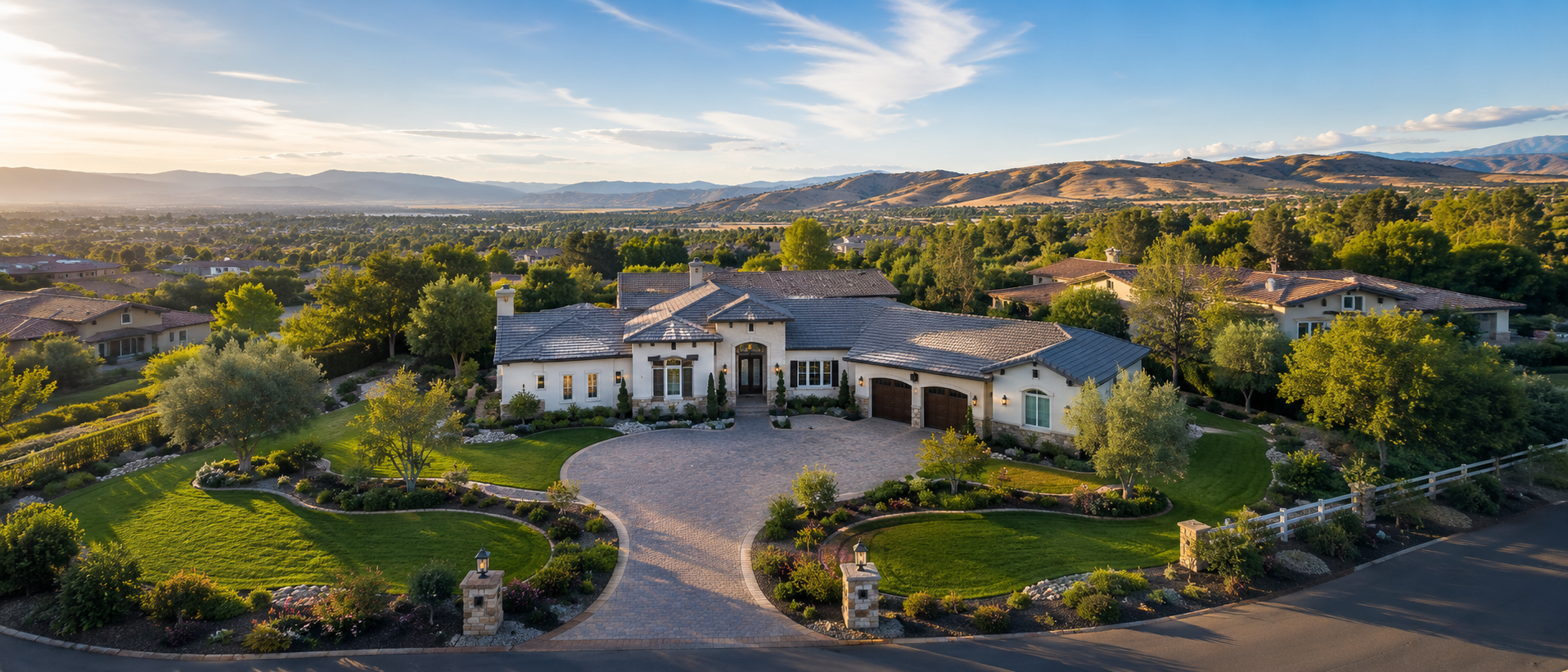 A large, luxurious house with a well-maintained front yard and driveway, set in a suburban neighborhood with other houses, lush trees, and a mountain range in the background under a partly cloudy sky.