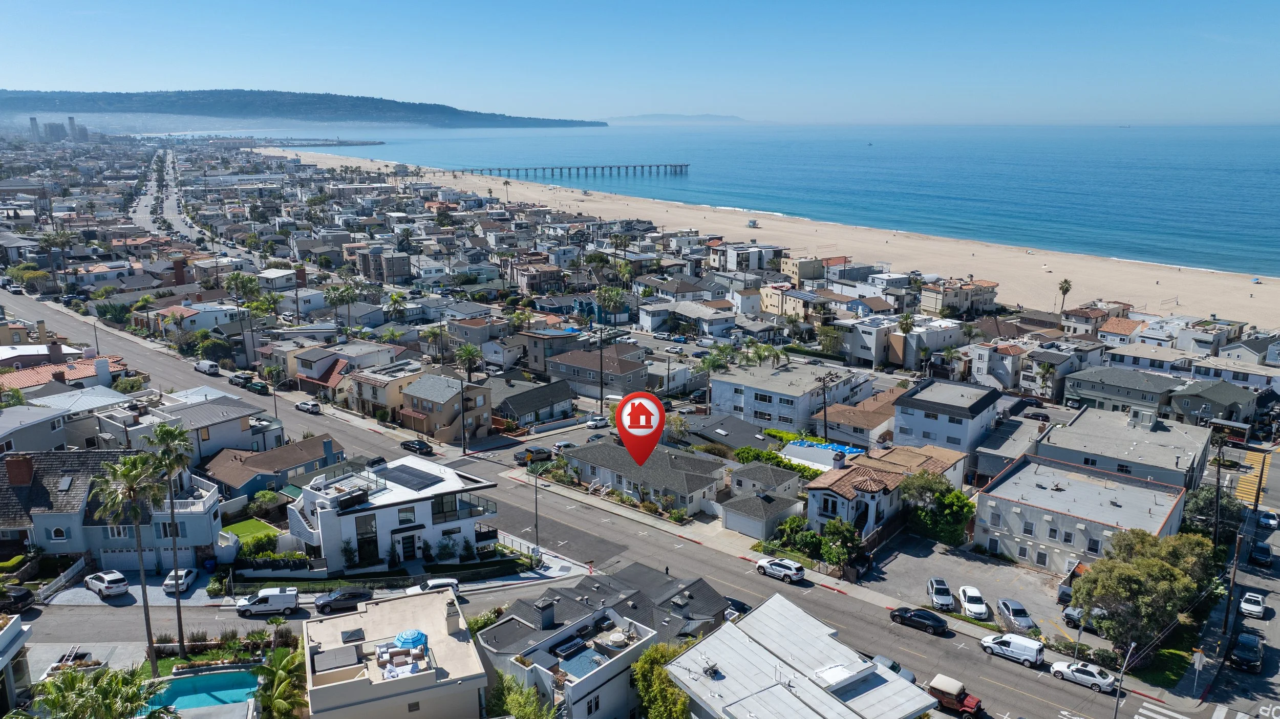 An aerial view of a coastal city with a beach and ocean on the right, lined with houses and streets. A pier extends into the water in the distance.