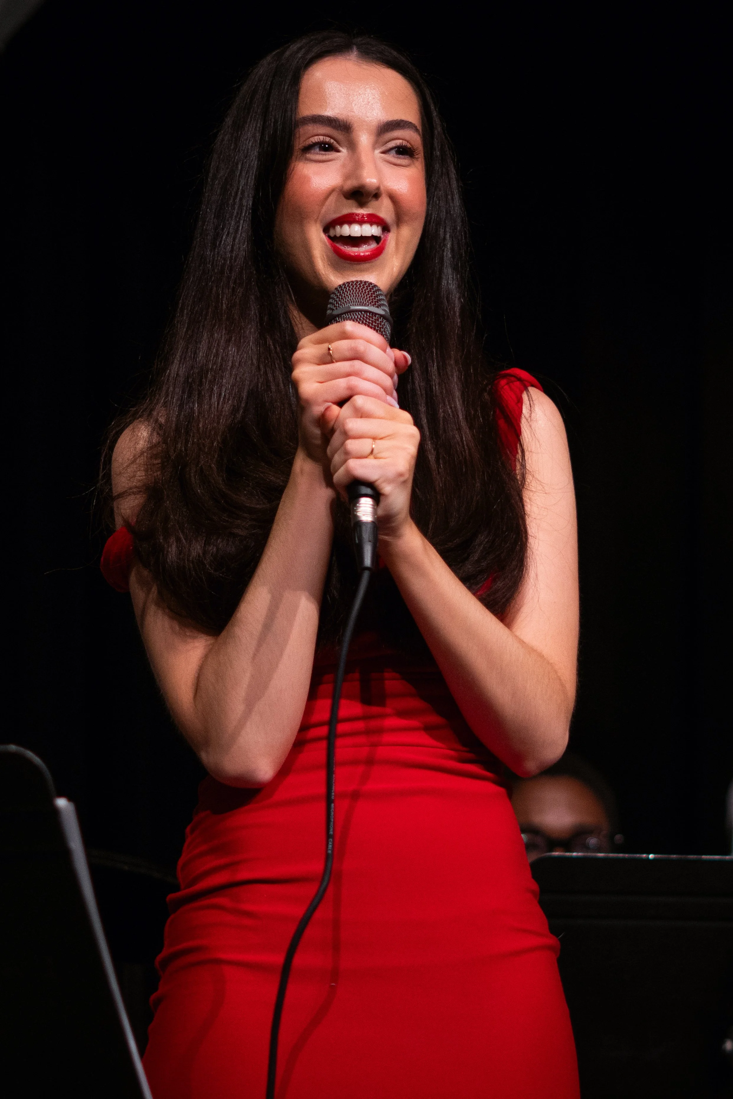 Woman in a red dress holding a microphone on stage, smiling.