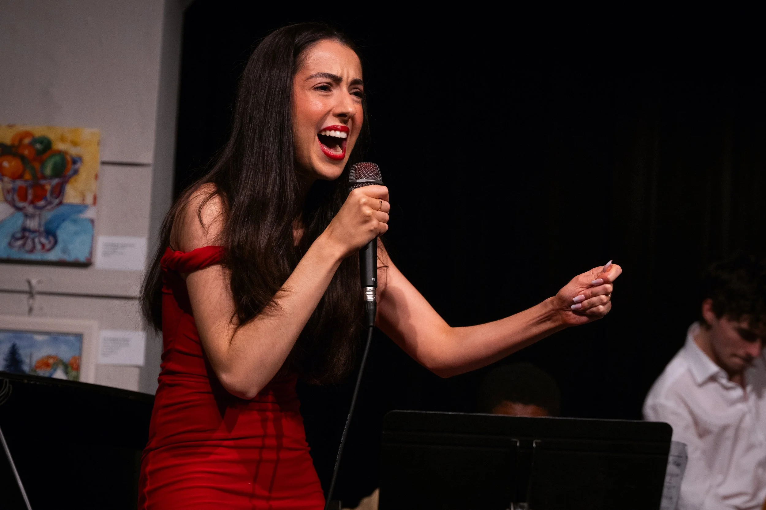 Woman in red dress singing passionately on stage, holding a microphone, with musicians in the background and artwork on the wall.