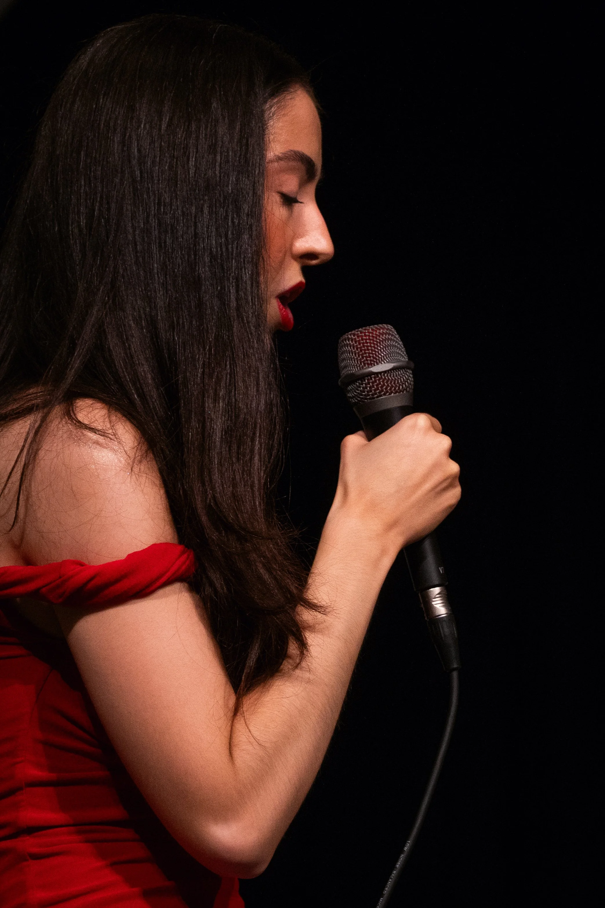 Woman in red dress singing into a microphone on stage, eyes closed, black background.