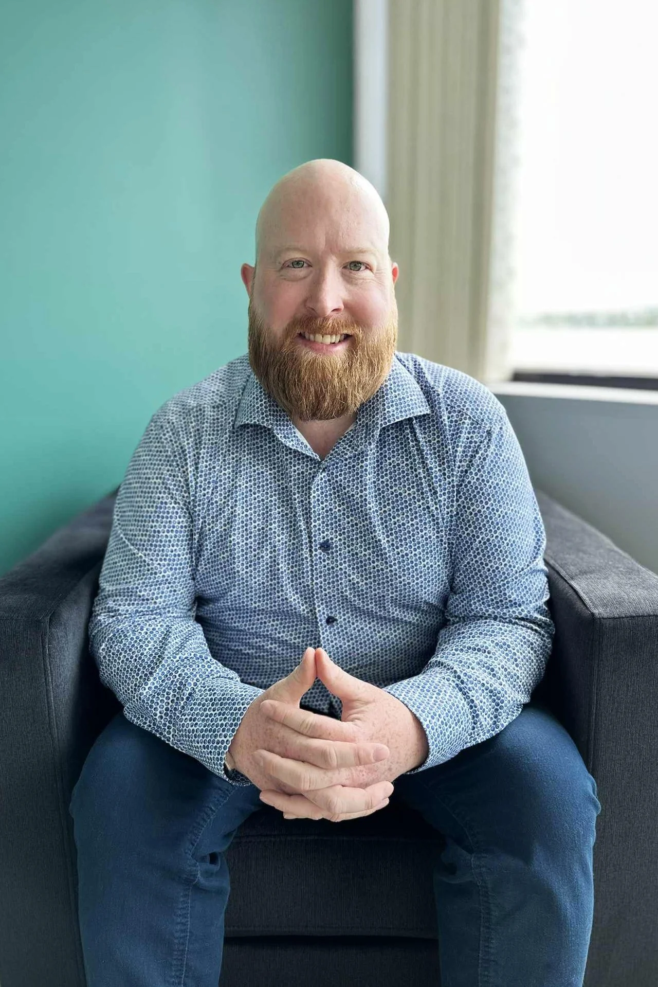 Man with a beard wearing a patterned dress shirt sitting on a dark gray sofa by a window, smiling with hands clasped.