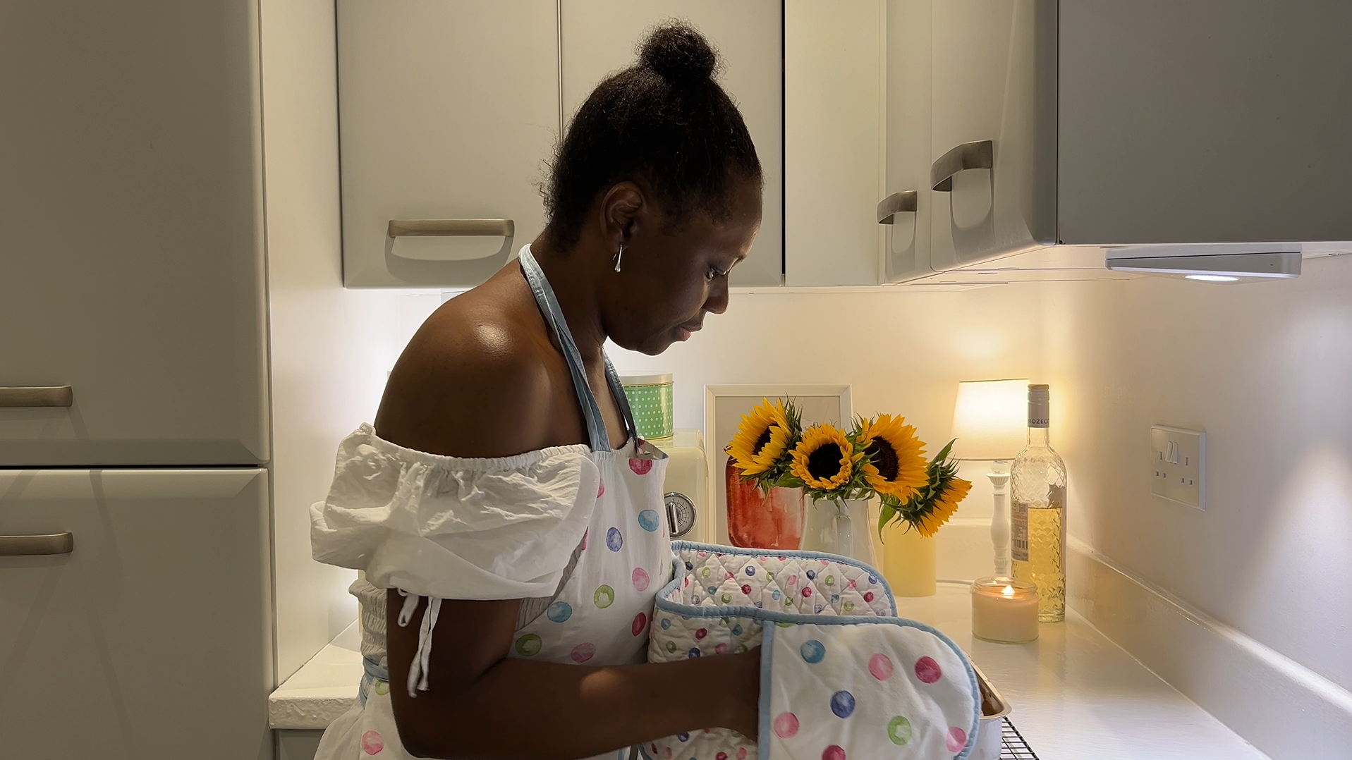 A woman wearing an apron and oven gloves holds a freshly baked dish in a warmly lit kitchen decorated with sunflowers and a candle.
