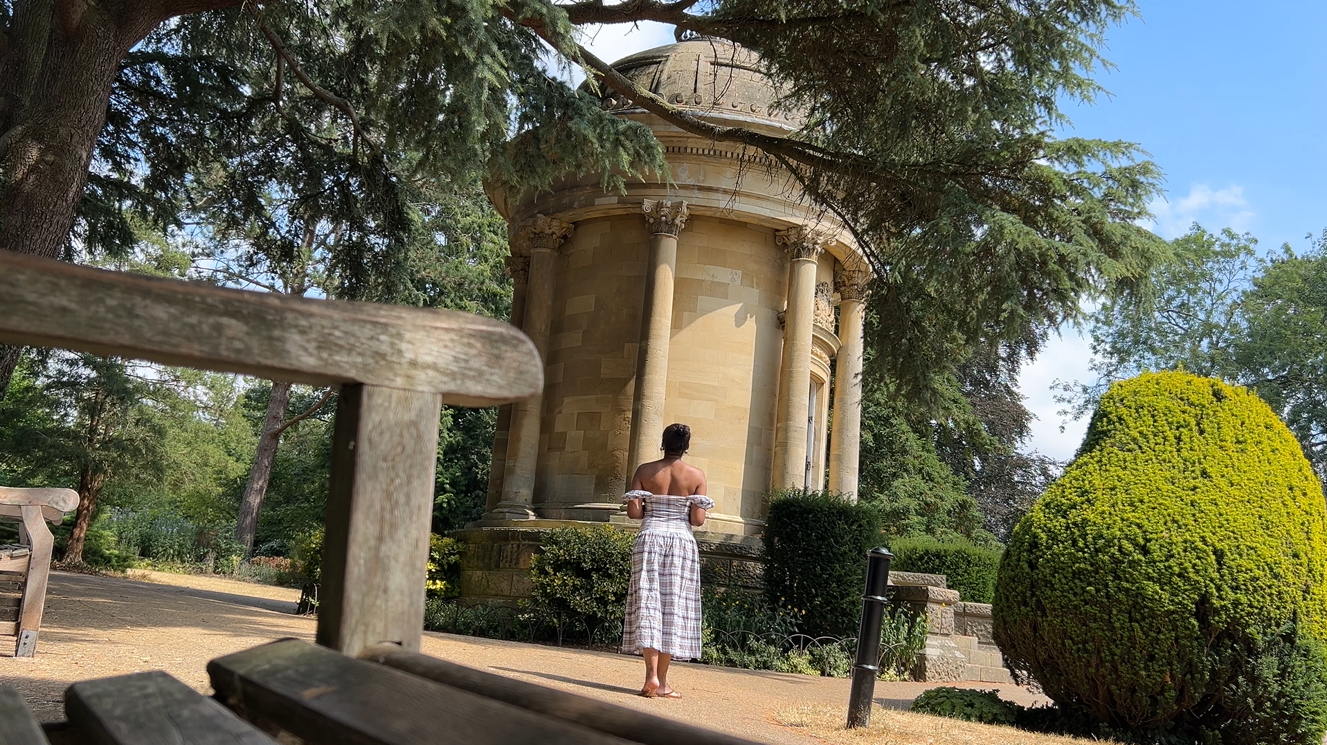 Eliane Luella stands in a checked summer dress beside a stone rotunda in a sunny park surrounded by trees and sculpted greenery.