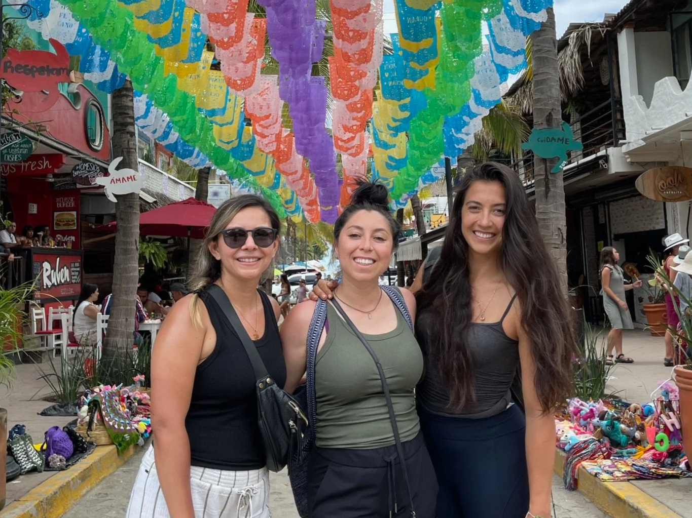 Three women smiling and standing close together on a busy street decorated with colorful papel picado banners. Street vendors with handmade crafts and souvenirs are visible in the background.