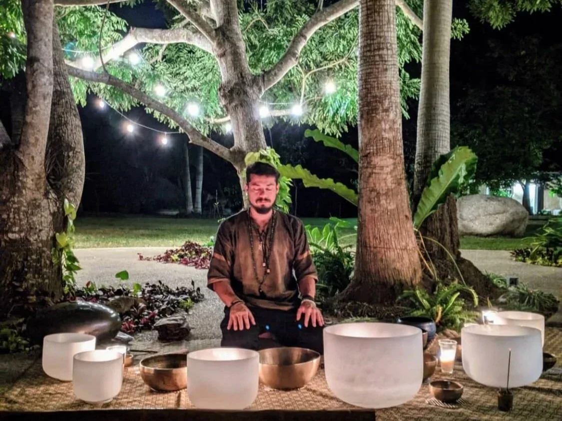 A man sitting outdoors at night with singing bowls in front of him, surrounded by trees and illuminated by string lights.