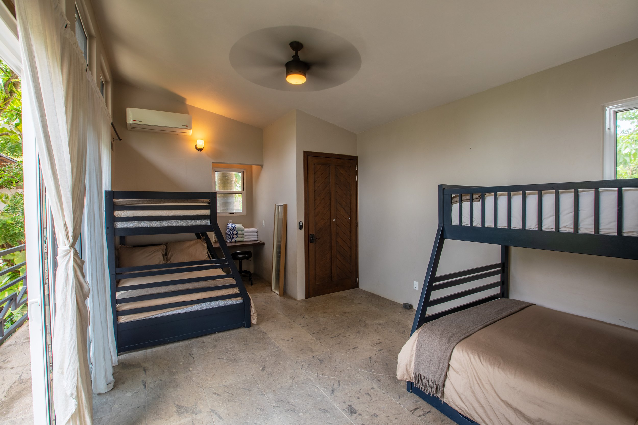 A room with two bunk beds with black frames, each with a mattress, a blanket, and pillows. There is a sliding glass door with curtains on the left side leading to a balcony or outside area. The room has a small window, a wooden door, a ceiling fan, an air conditioner, and a light fixture on the wall. There are some folded towels on a desk near the window, and the floor is made of stone or tile.