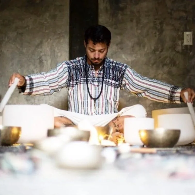 A man with dark hair and a beard, wearing a striped kurta, sits cross-legged with eyes closed, performing a ritual surrounded by bowls and offerings on the ground against a textured wall.