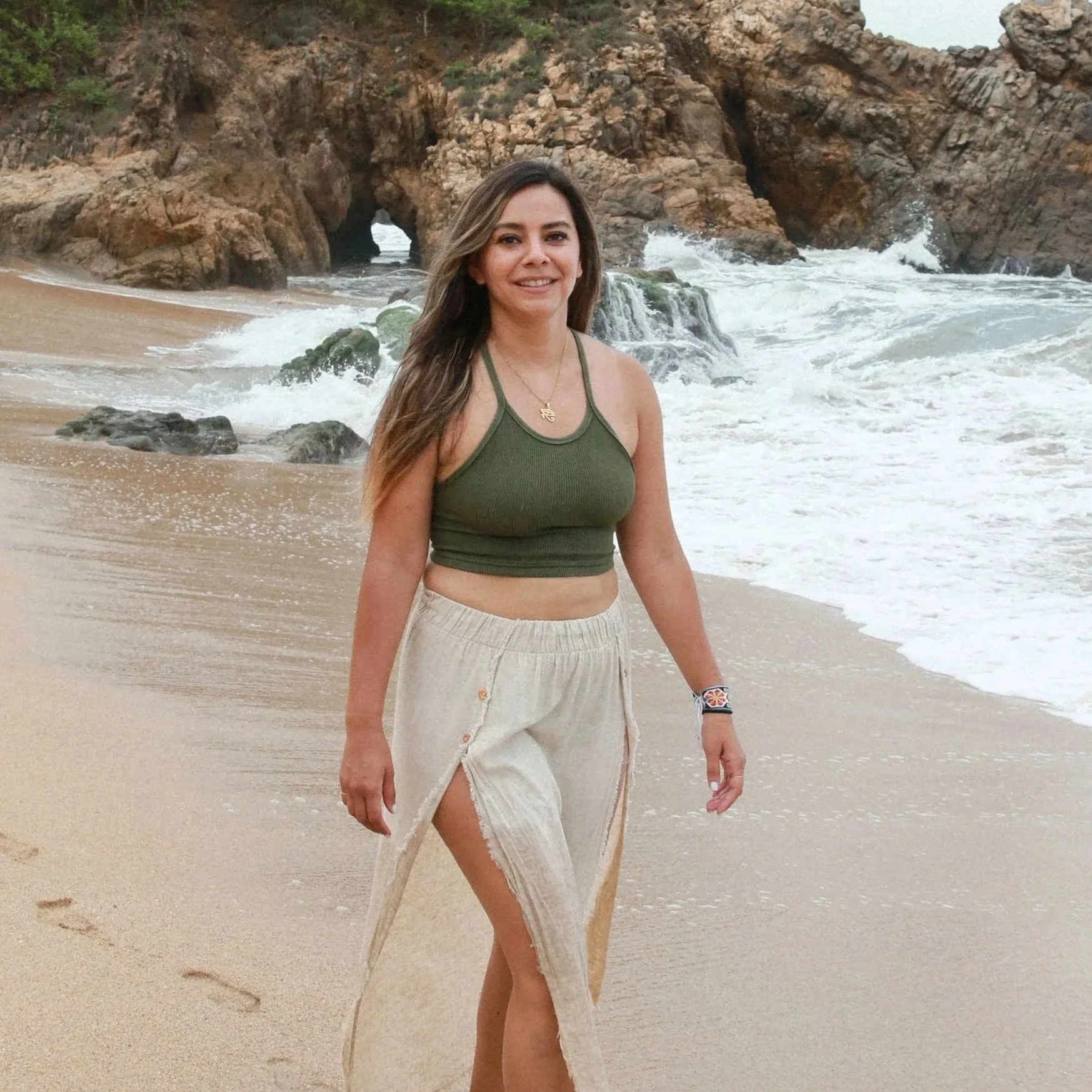A woman smiling on a beach with rocky cliffs and ocean waves in the background.