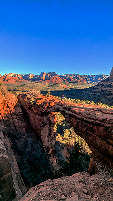 A person standing on Devil's Bridge, elevated rock formation in a desert canyon, surrounded by red rock cliffs and green vegetation under a clear blue sky.