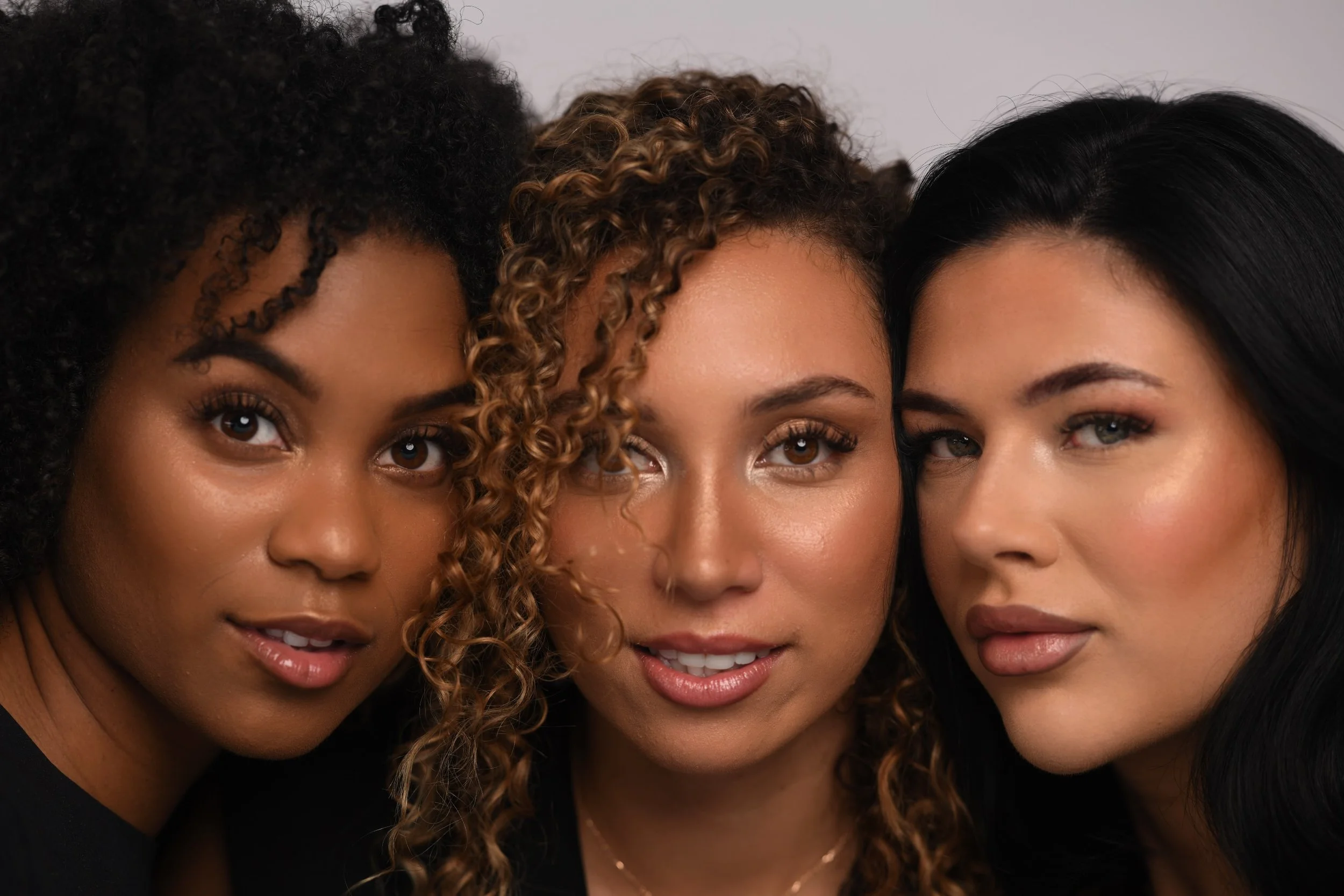 Close-up portrait of three women with diverse hairstyles and makeup, looking confidently at the camera against a neutral background.