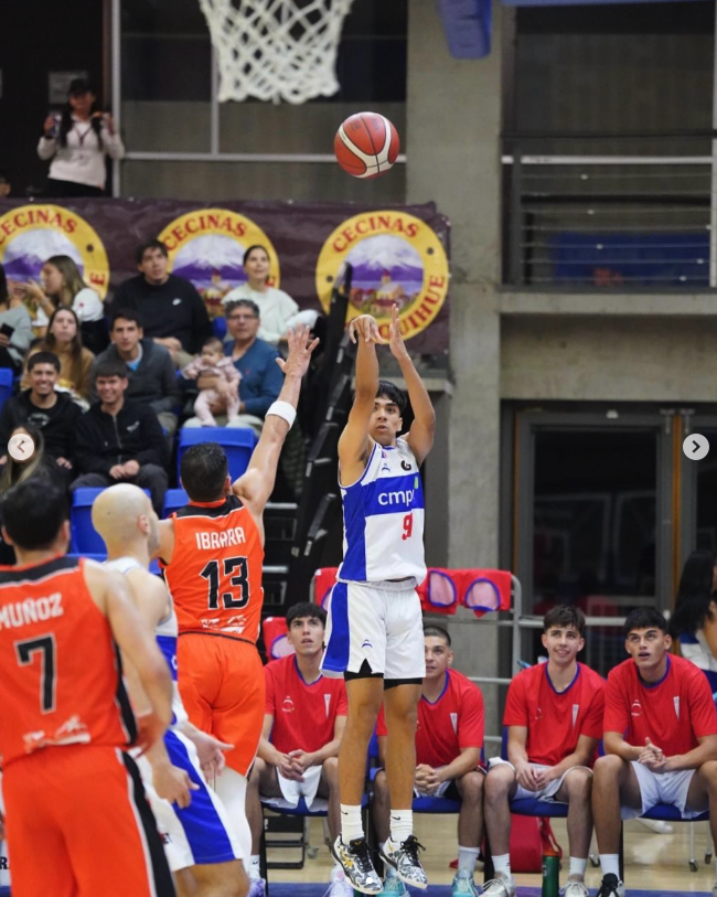 Juego de baloncesto con jugadores en acción en una cancha cubierta, espectadores en el fondo y un equipo sentado en el banquillo.