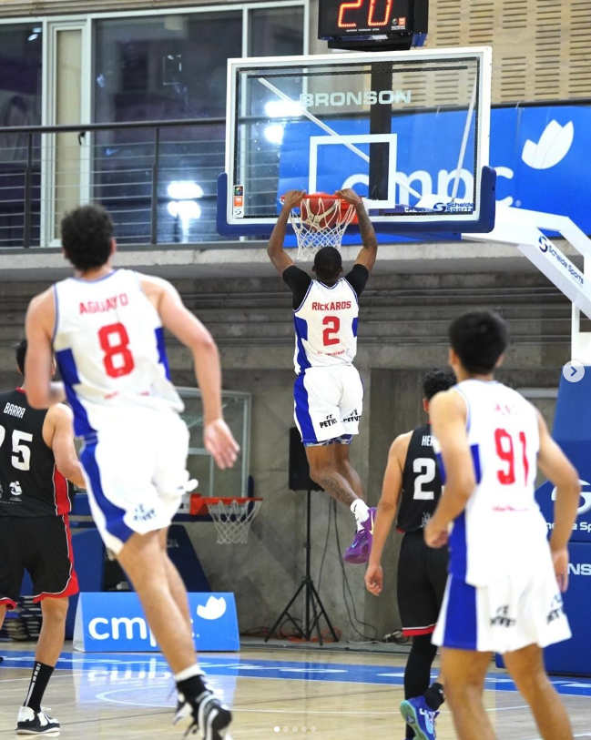 Jugador de baloncesto número 2 con uniforme blanco y azul, saltando para encestar, en un partido en interiores. Otros jugadores en el suelo, algunos en uniforme negro y otros en blanco, observan. Pantalla digital y anuncios en el fondo.
