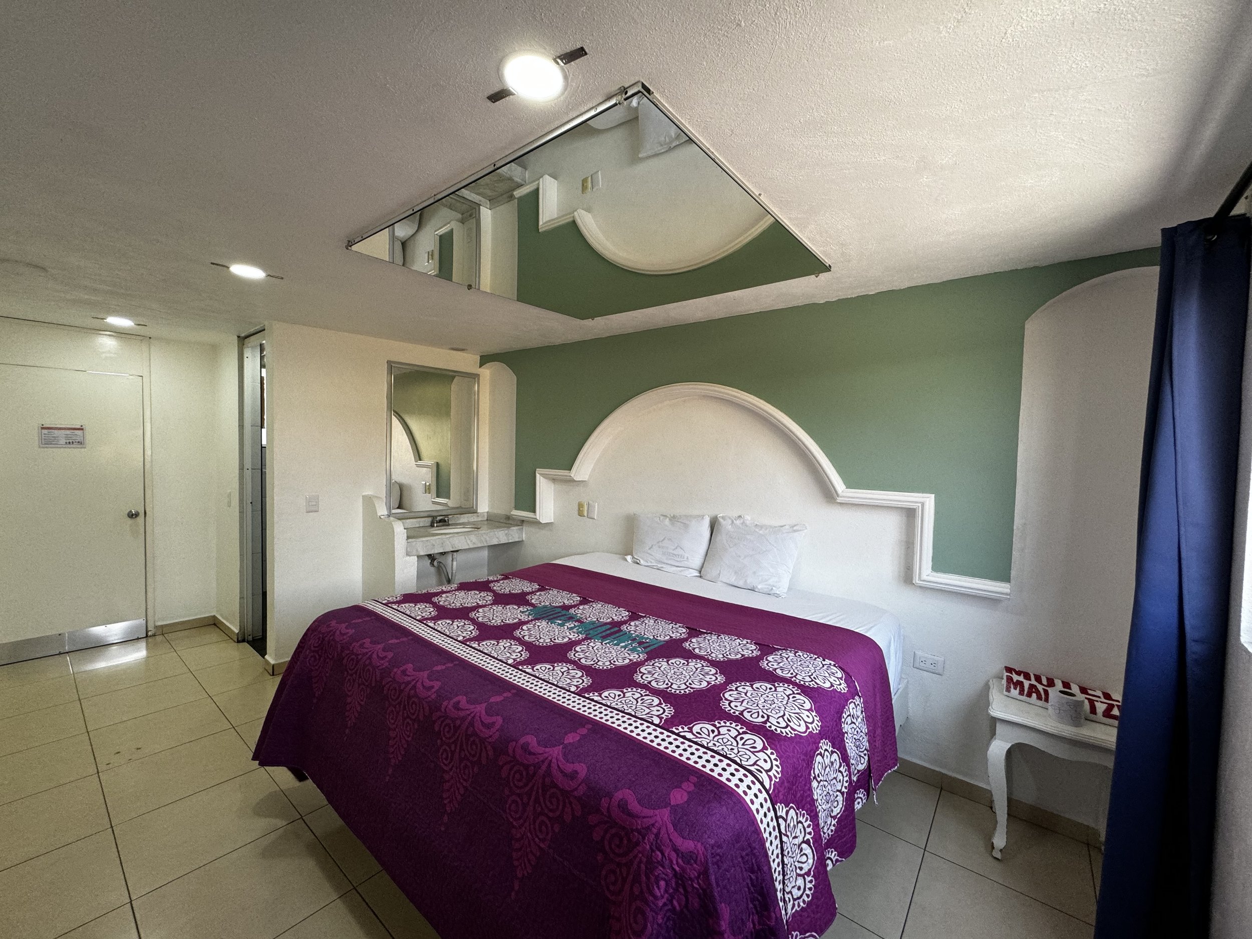 Habitación de hotel con cama doble, colcha morada con diseño blanco, espejo en el techo, mesa de noche blanca, y pared verde claro. La habitación tiene una pequeña área de tocador con un espejo y está bien iluminada.