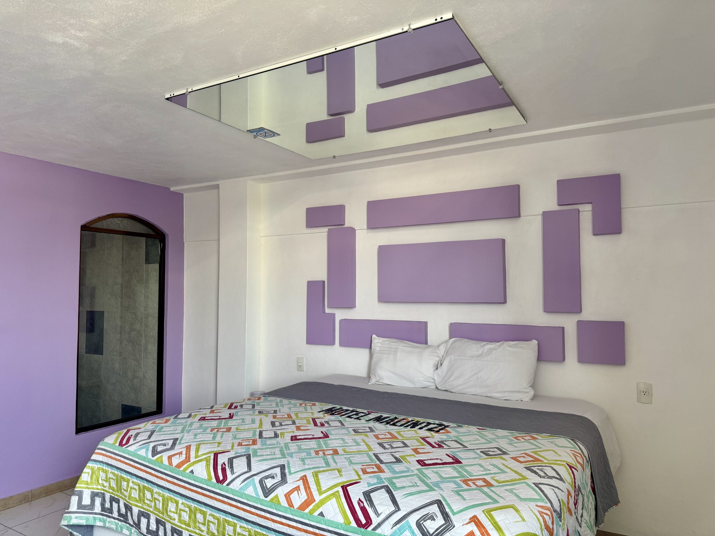 Habitación con cama, decoración geométrica morada en la pared, edredón colorido, espejo en el techo y pared pintada de morado.