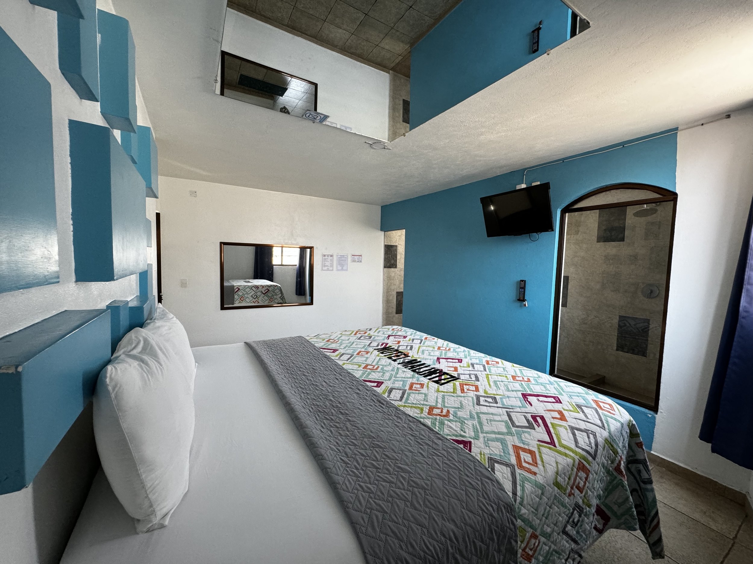 Habitación con cama grande, colcha colorida, almohadas blancas, paredes azules, televisor montado en la pared, espejo en el techo y ventana pequeña. Baño visible a través de una puerta con arco.