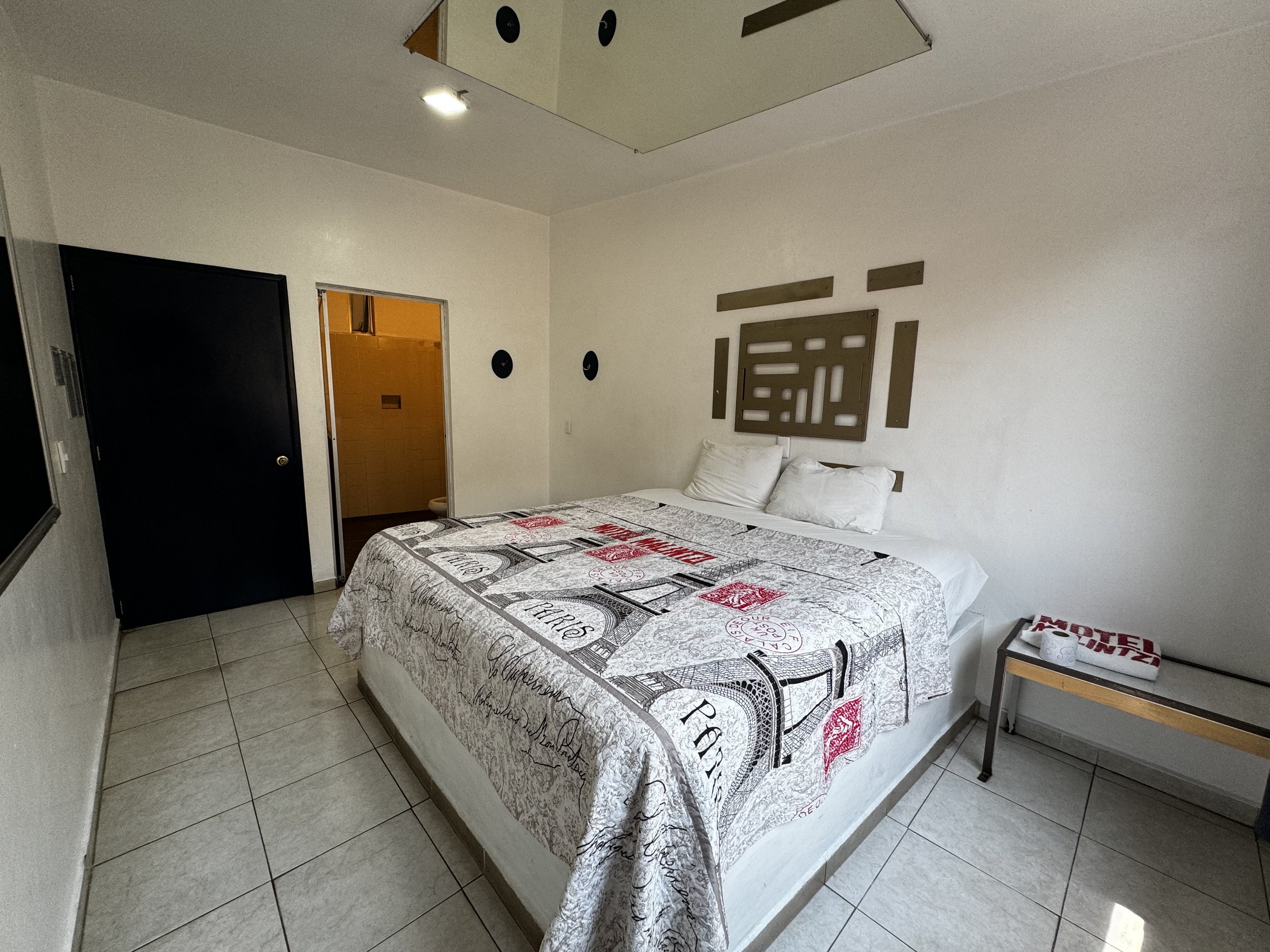Habitación de hotel con cama grande, paredes blancas, edredón estampado, decoración en la pared, puerta negra y pequeño baño visible a la izquierda.