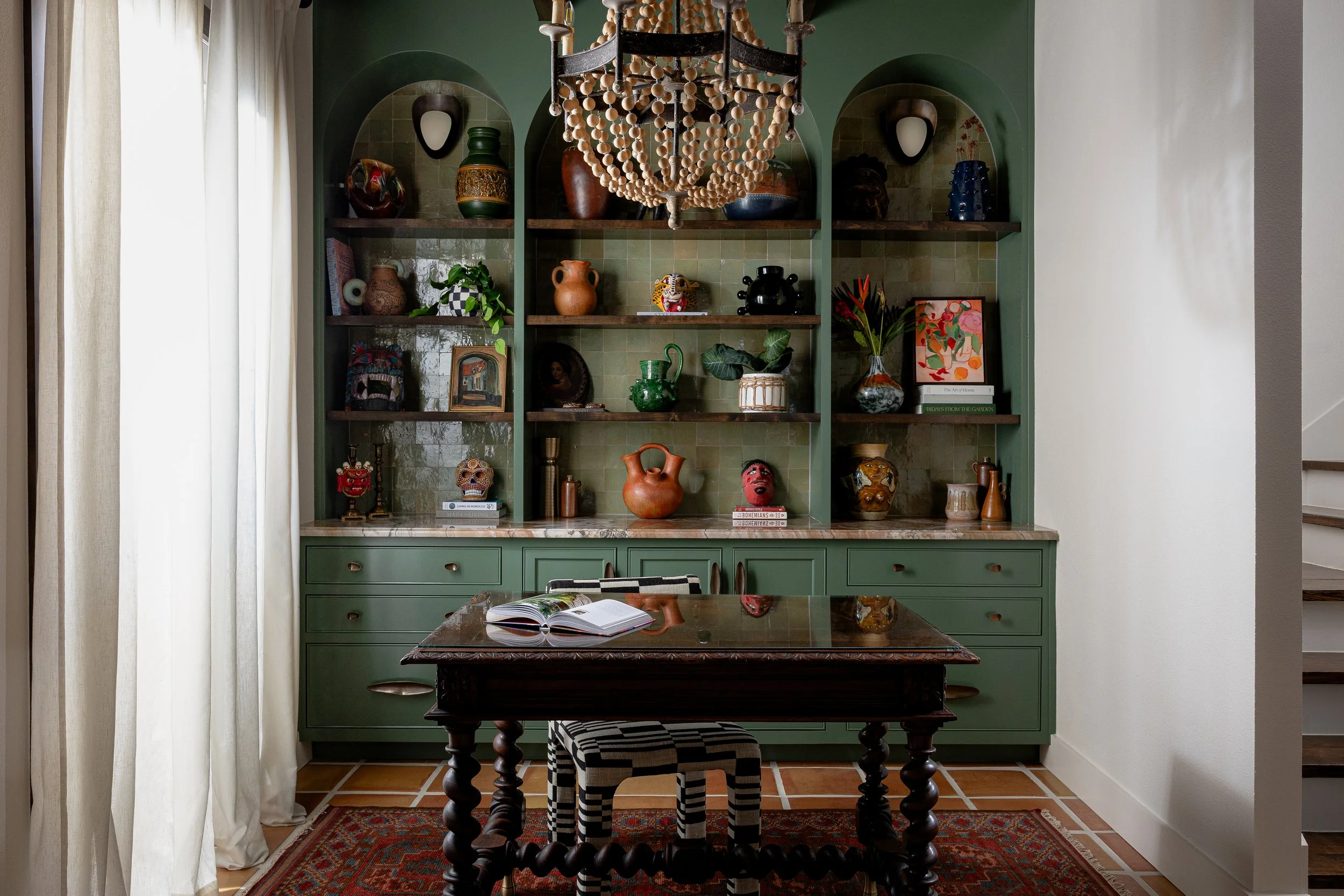 A dining room featuring a green built-in cabinet with open shelves displaying colorful vases, artwork, and decorative items. A wooden dining table with an open book and black-and-white checkered stool is in the foreground. A large beaded chandelier hangs above the table, and natural light filters in through sheer curtains on the left.