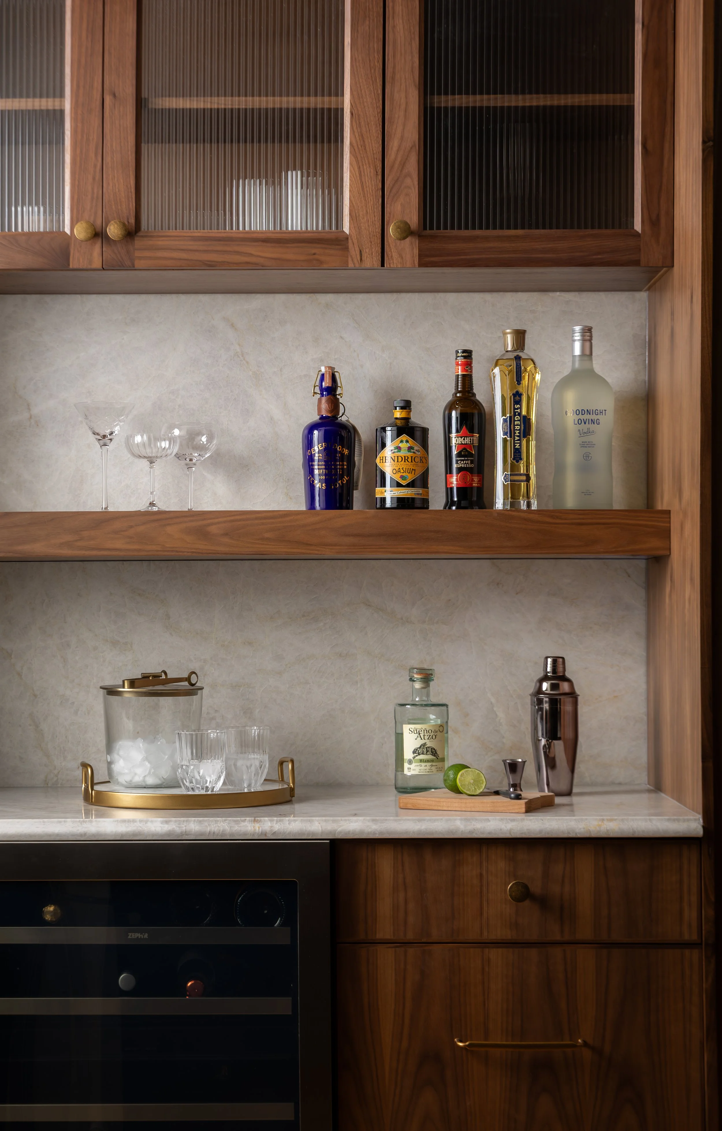 Kitchen shelf with liquor bottles, three glasses, ice bucket, and lime slices on a marble countertop.