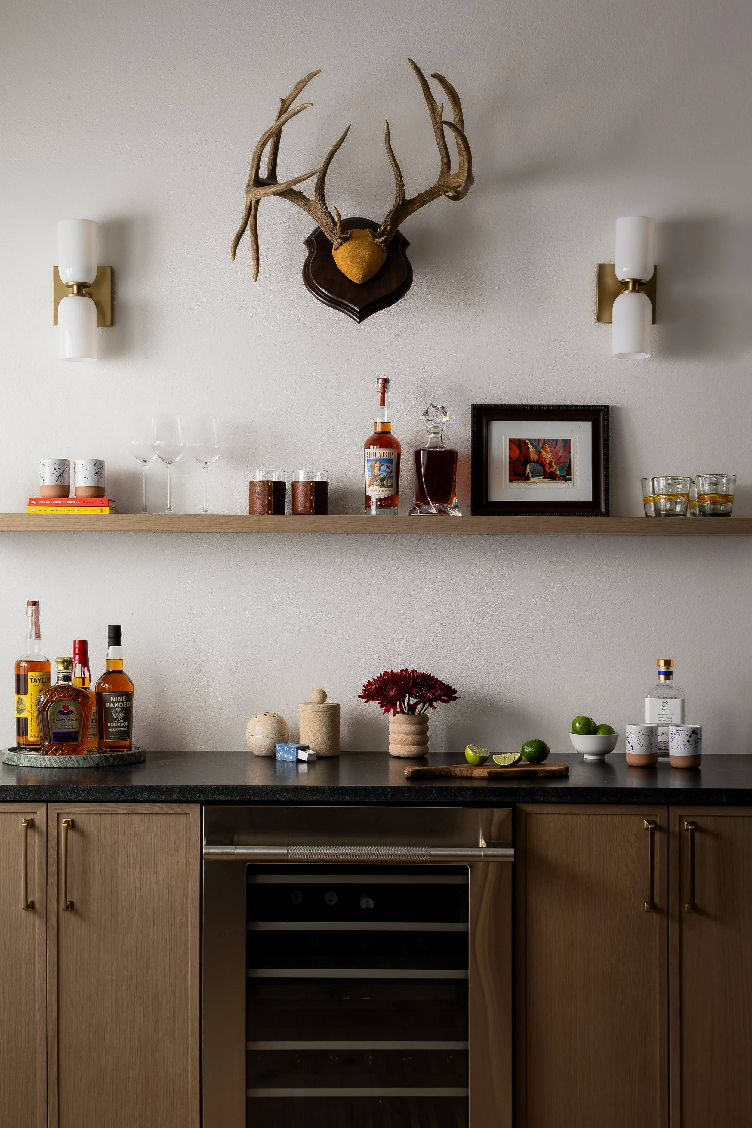 A kitchen counter with various bottles of alcohol, a bunch of limes, and small jars, with a beige cabinet below. Above, a wooden shelf holds decorative items and a framed picture. Mounted on the wall are antlers with a mounted deer's head and two wal