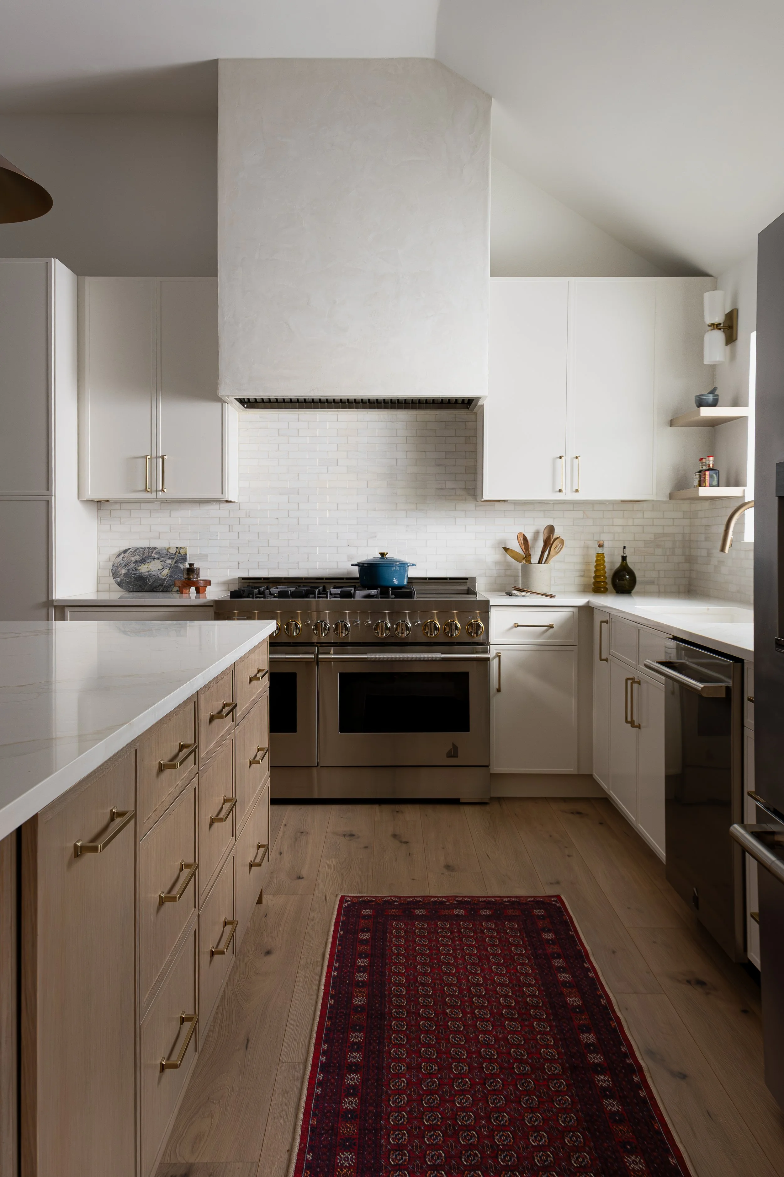 A modern kitchen with white cabinets, a stainless steel stove, a white brick backsplash, wooden flooring, and a red patterned rug.