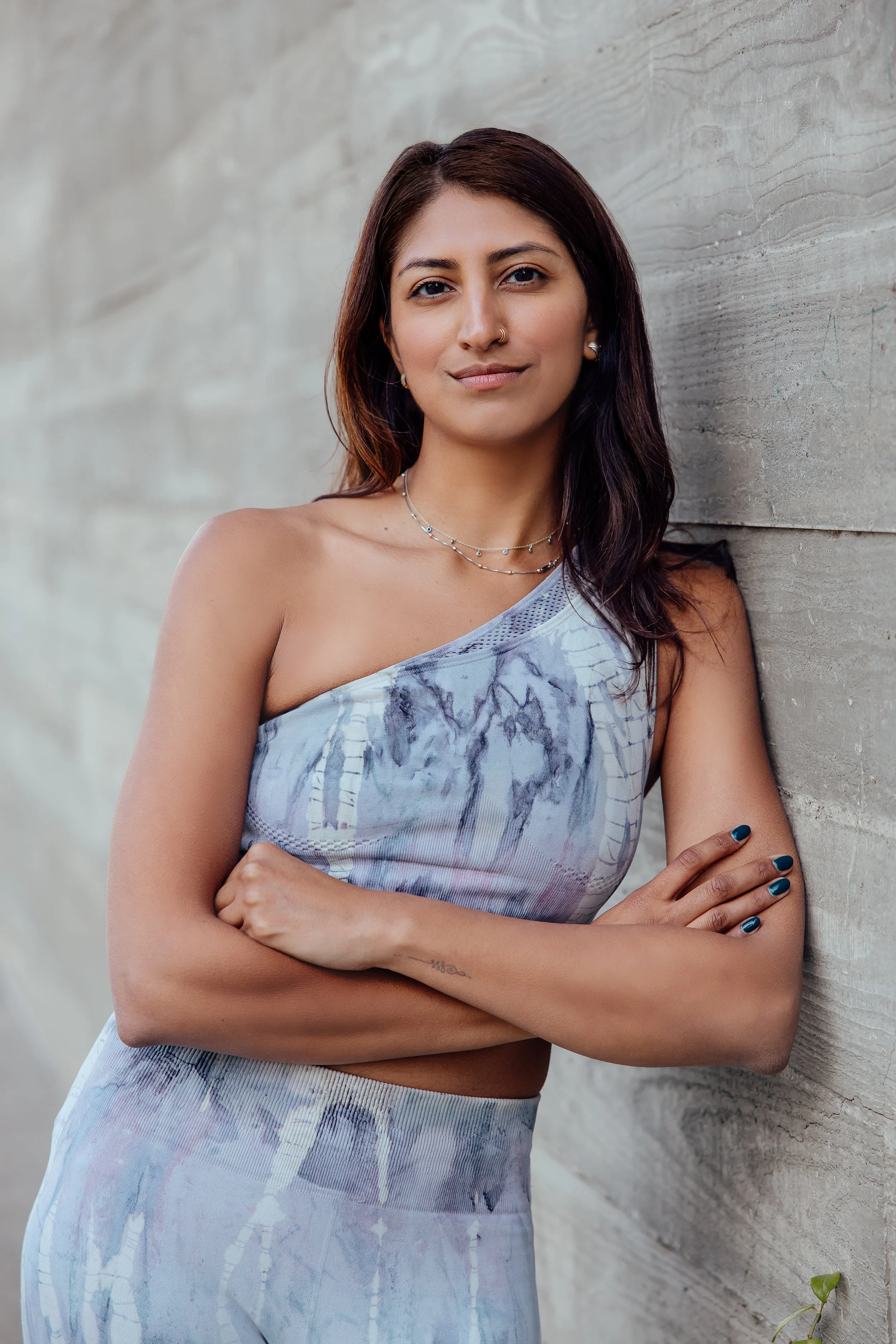 Young woman with dark brown hair standing against a concrete wall, wearing a sleeveless, patterned workout outfit and jewelry.