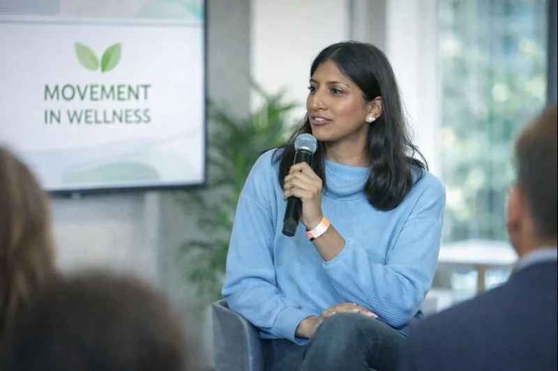 A woman with black hair in a blue sweater speaking into a microphone at a wellness event, with a screen displaying 'Movement in Wellness' in the background.