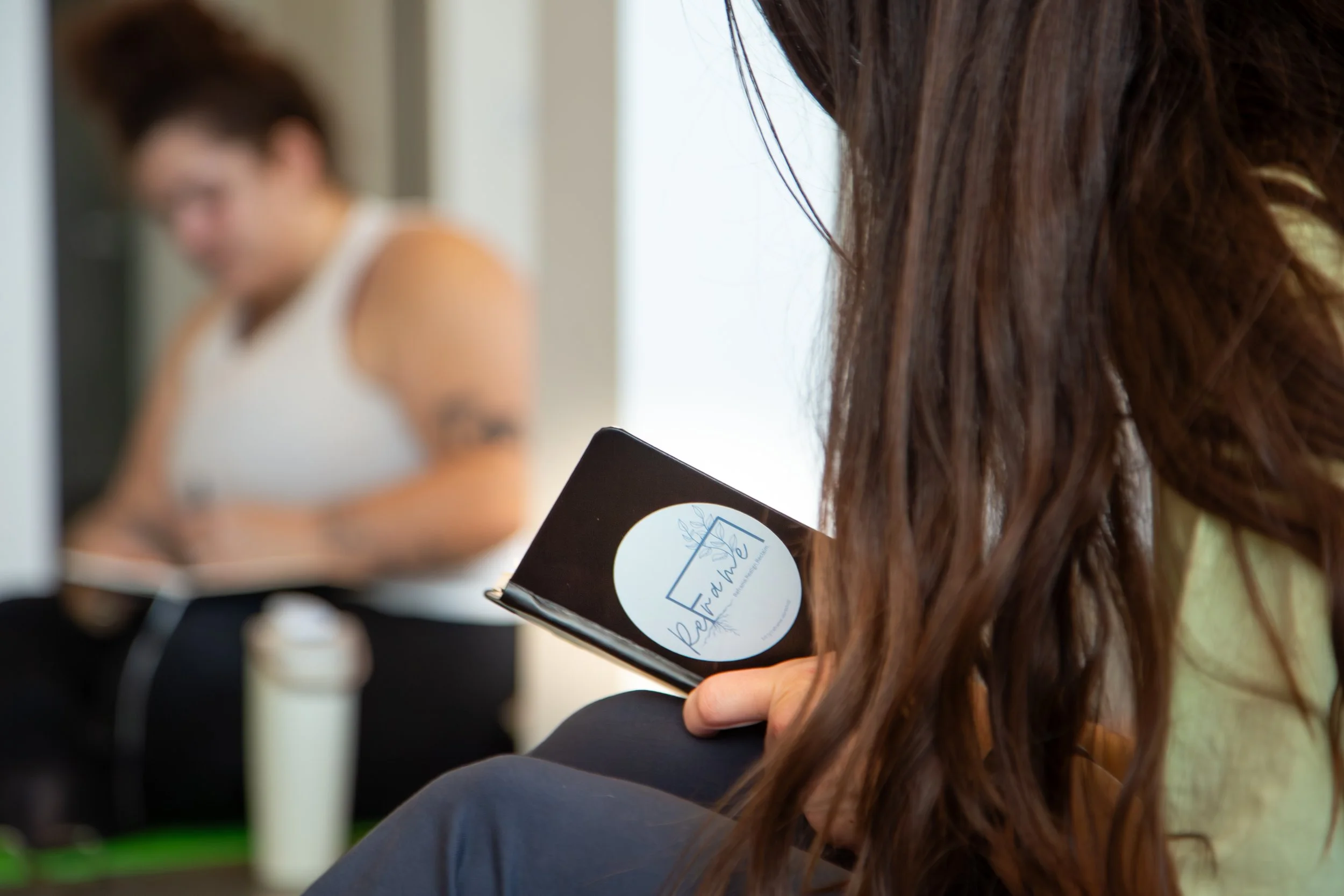 A woman with long brown hair looking at her phone with a sticker that says "ReWilded" with a mountain and leaf logo, while another woman in a white tank top is blurred in the background.
