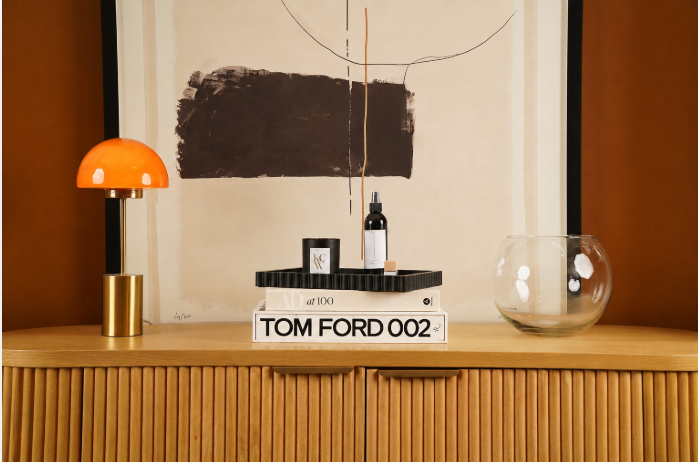 Decorative wooden sideboard with a small orange and gold lamp, art books, a small bottle, and a glass bowl on top, in front of a modern abstract wall art.