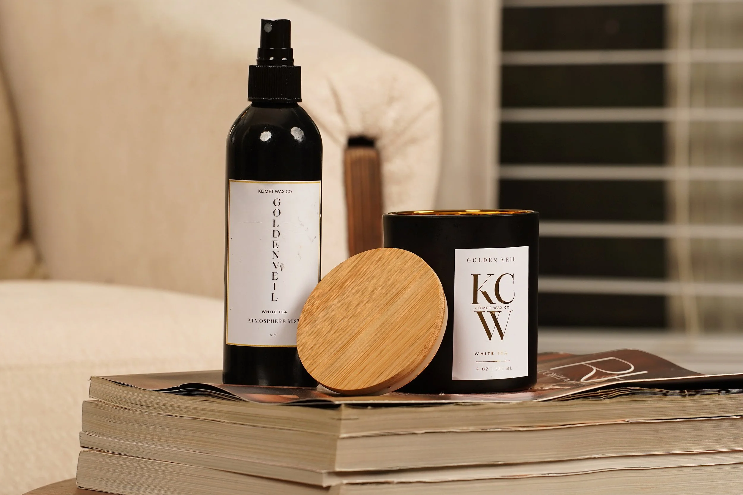 A black spray bottle, a black candle, and a round wooden object are arranged on a stack of magazines on a table.