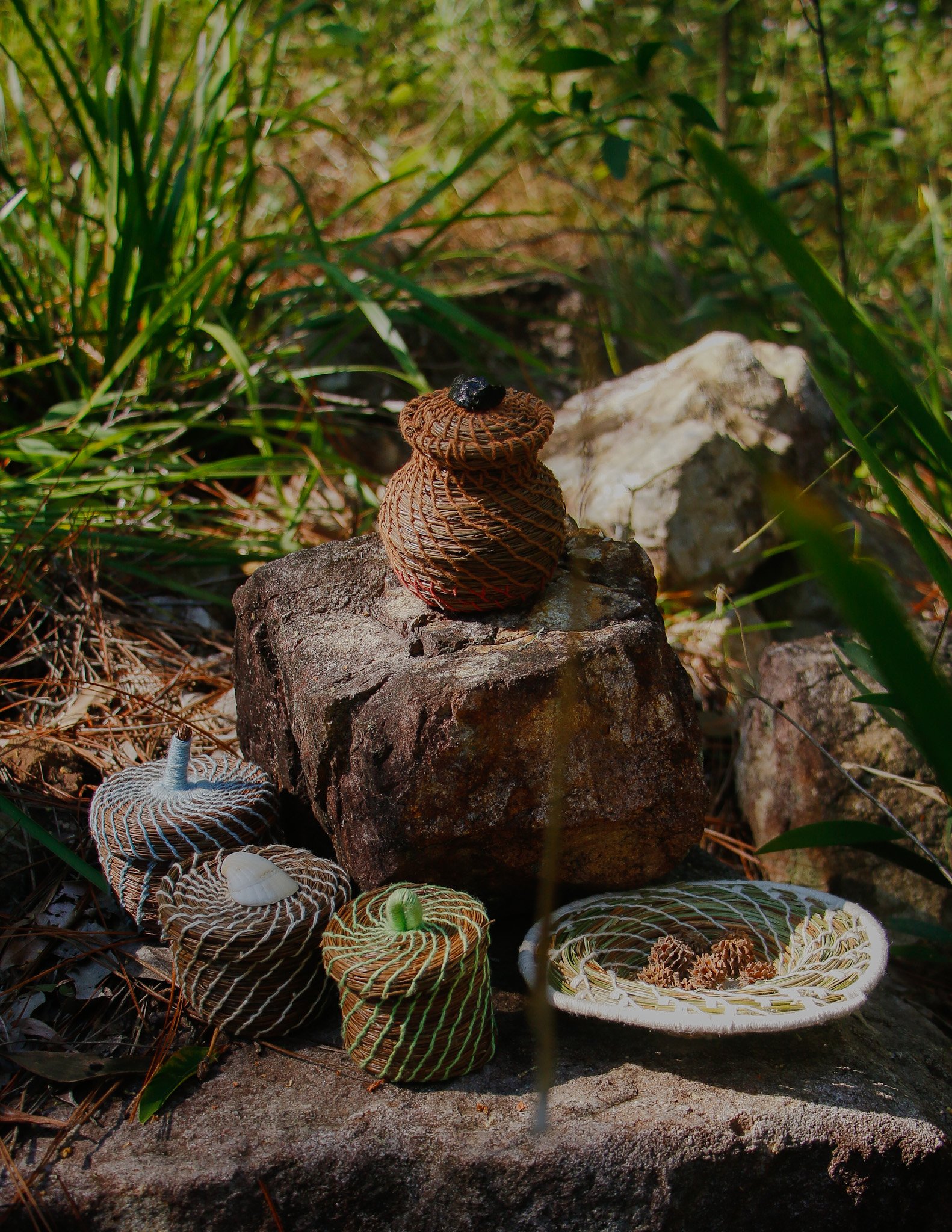 Colorful handmade woven baskets on rocks and ground in outdoor setting with grass and plants.