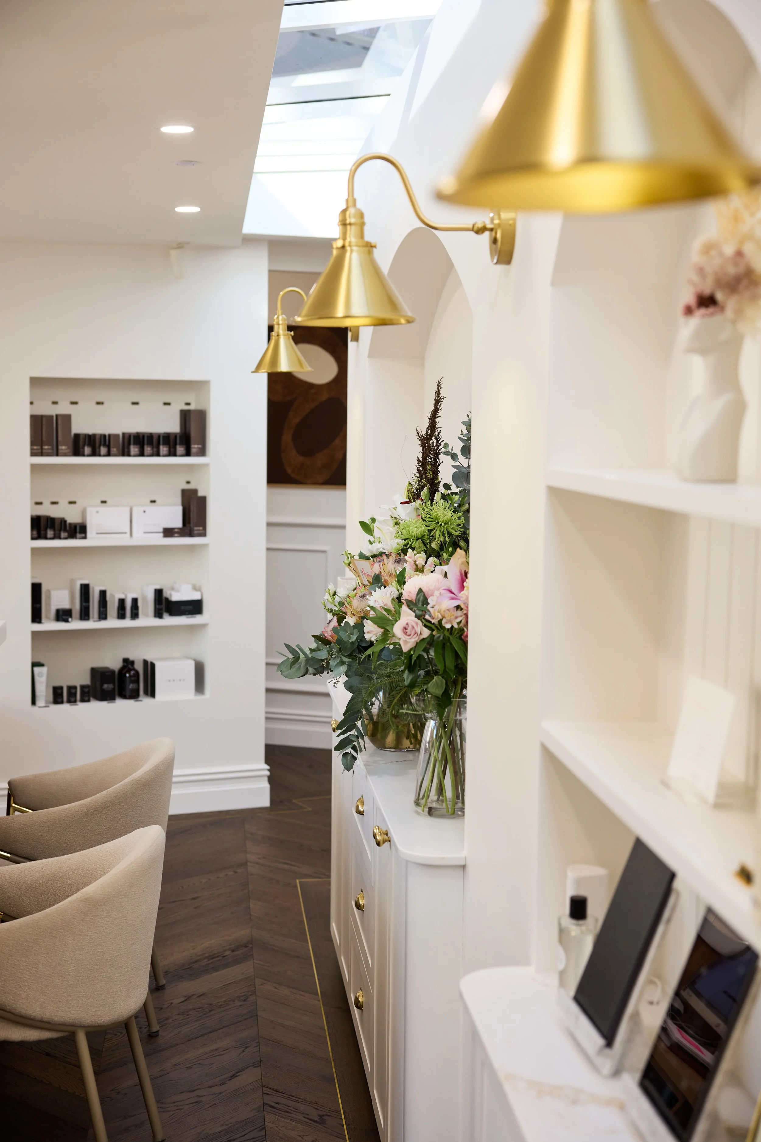 Interior of a modern space with white walls, gold wall-mounted lamps, a white cabinet with gold knobs, a large floral arrangement in a glass vase, and beige chairs with wooden legs, dark wooden flooring, and shelves with various products.