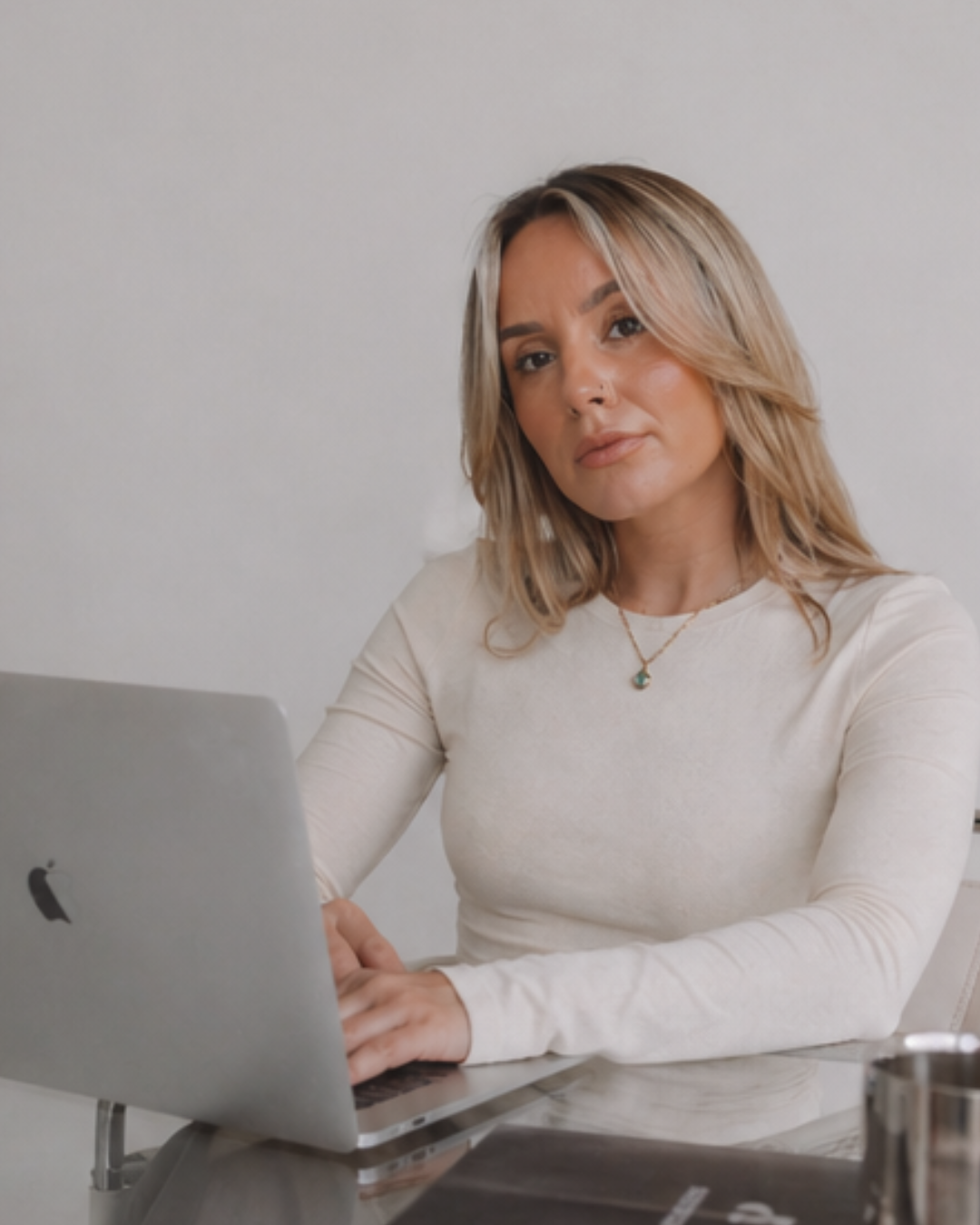 A woman with shoulder-length blonde hair working on a silver MacBook laptop at a glass table, wearing a cream long-sleeve top and a necklace, in a minimalistic room with a plain white wall background.