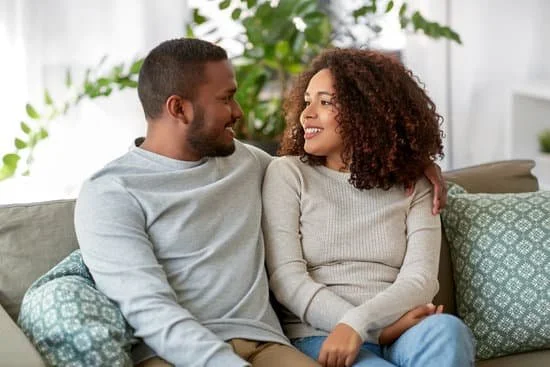 A couple sitting on a couch, smiling and looking at each other, in a bright living room with plants in the background.