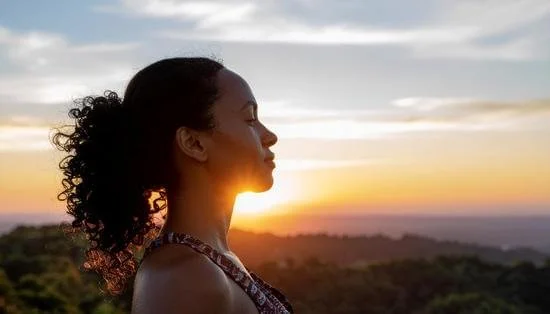 Profile of a woman with curly hair standing outdoors at sunset, with a landscape and sky in the background.