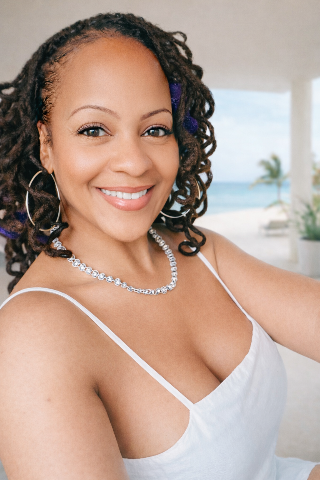 A woman smiling at the camera, wearing a white tank top, jewelry, and earrings, with a beach and ocean in the background.