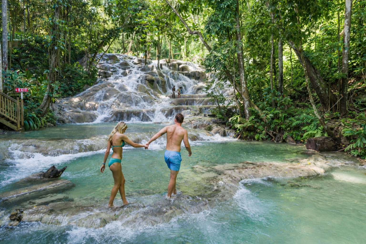 A couple holding hands while walking in a natural swimming hole surrounded by lush jungle, with a waterfall in the background.
