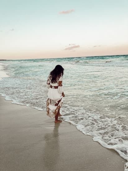 A woman with long dark hair walking along the shoreline of a beach during sunset, wearing a white dress with a floral pattern.