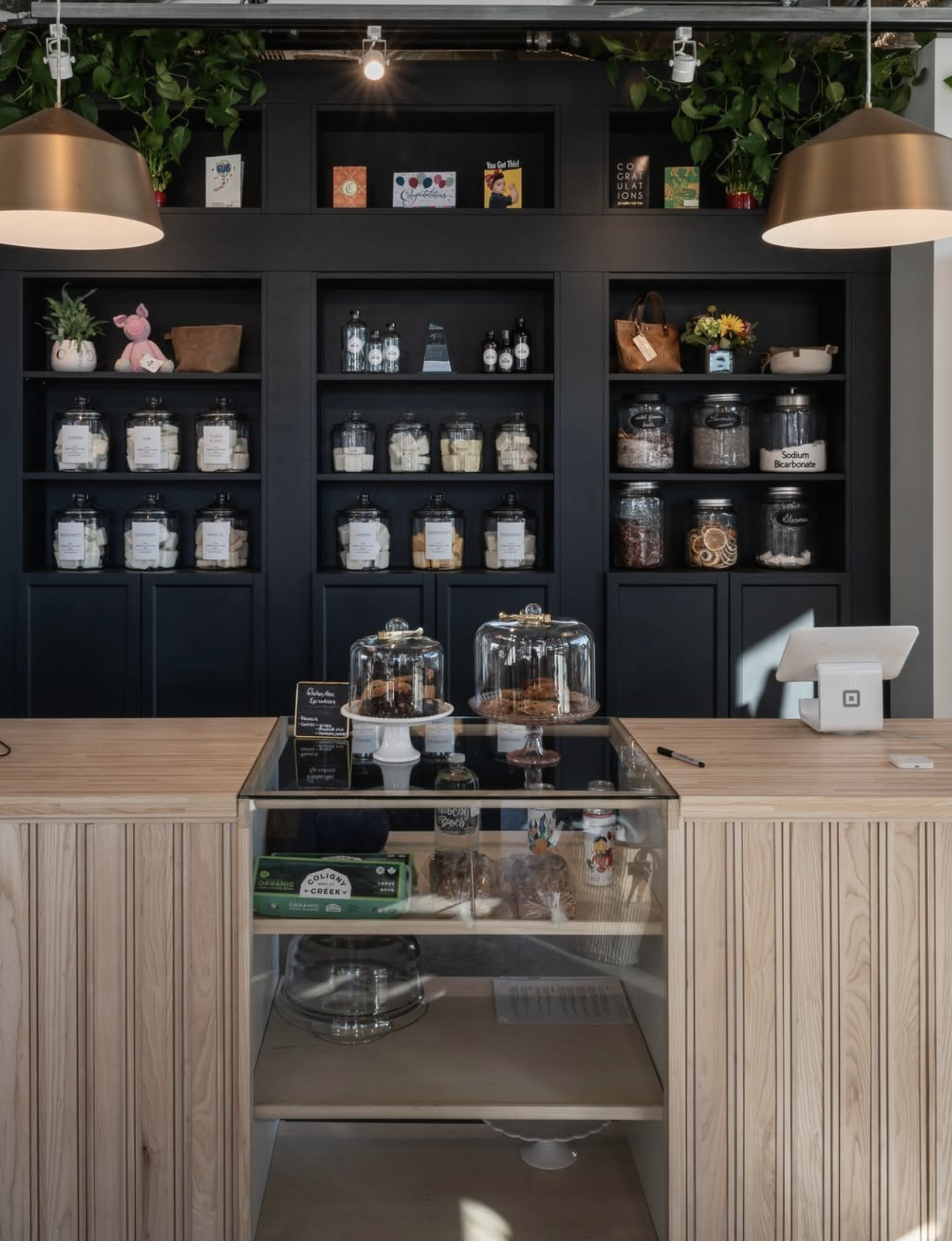 A modern bakery display with a wooden counter, glass cake domes with baked goods, and shelves filled with jars of ingredients, plants, and greeting cards in the background.
