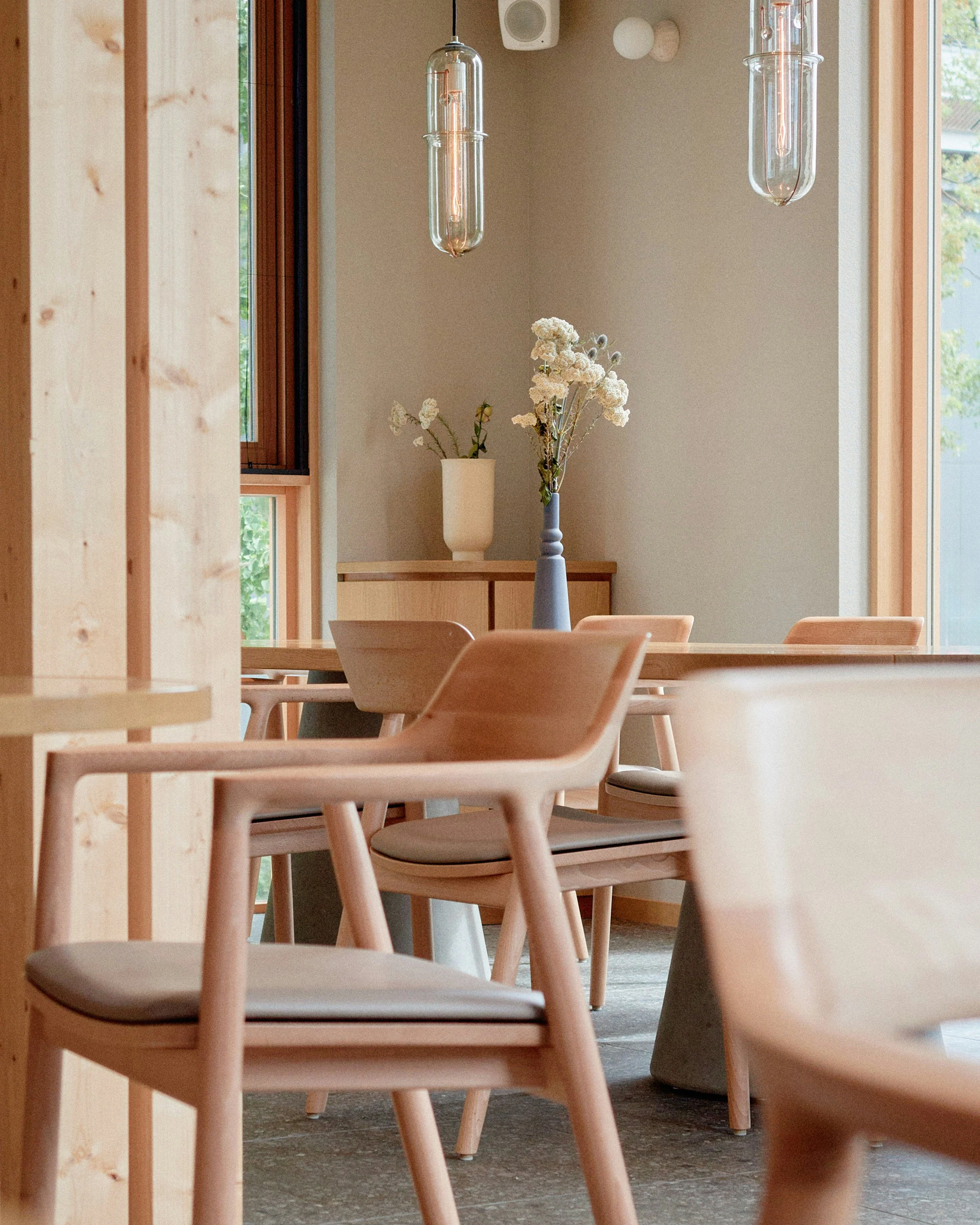 A dining area with wooden chairs around a table, decorated with a tall blue vase holding white flowers, and another white vase with green foliage on a sideboard. Stylish hanging glass light fixtures are above, with windows allowing natural light to enter.