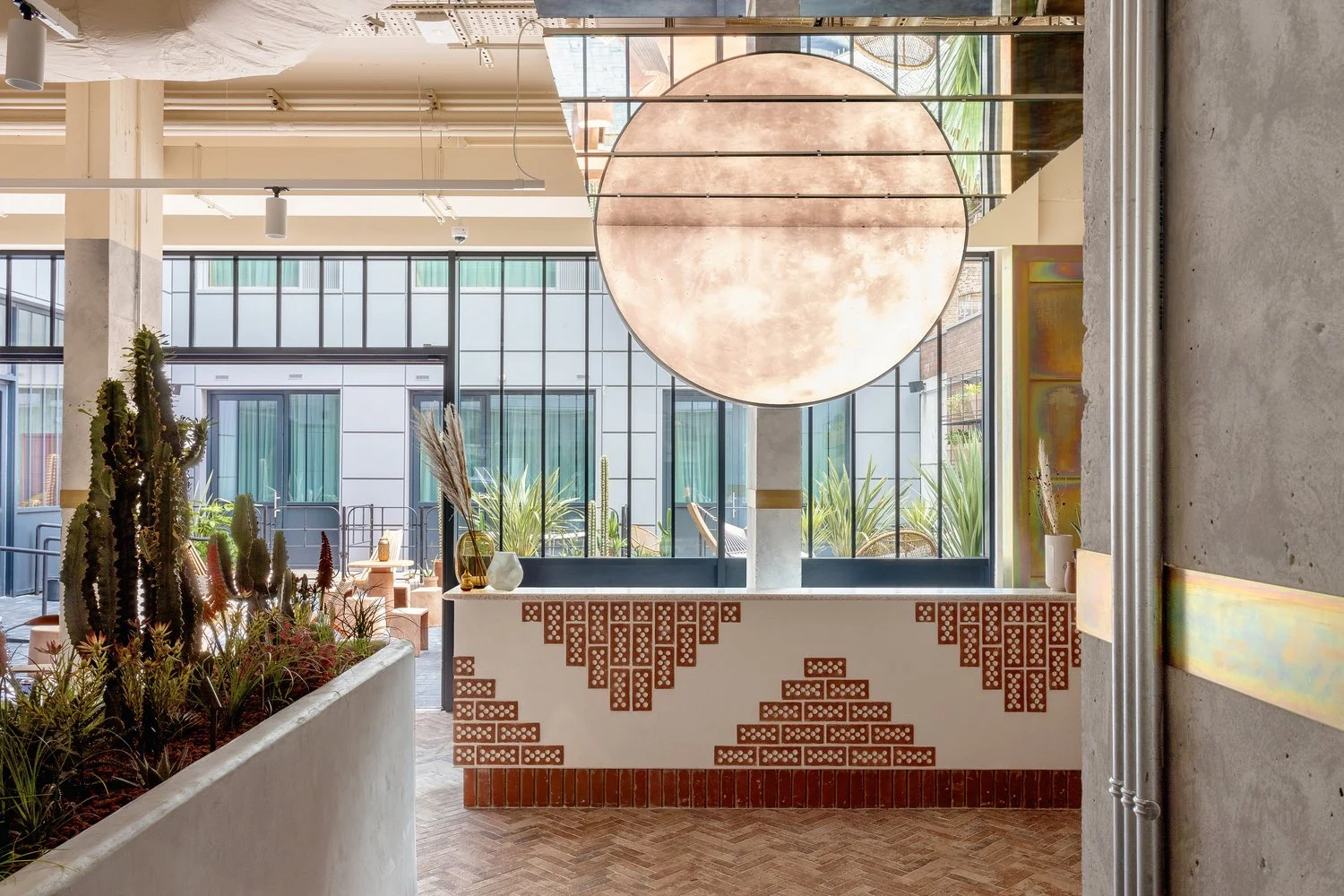 Interior of a modern hotel with a large round light fixture, cacti and plants, a white brick-patterned wall, and large glass windows showing outdoor plants.