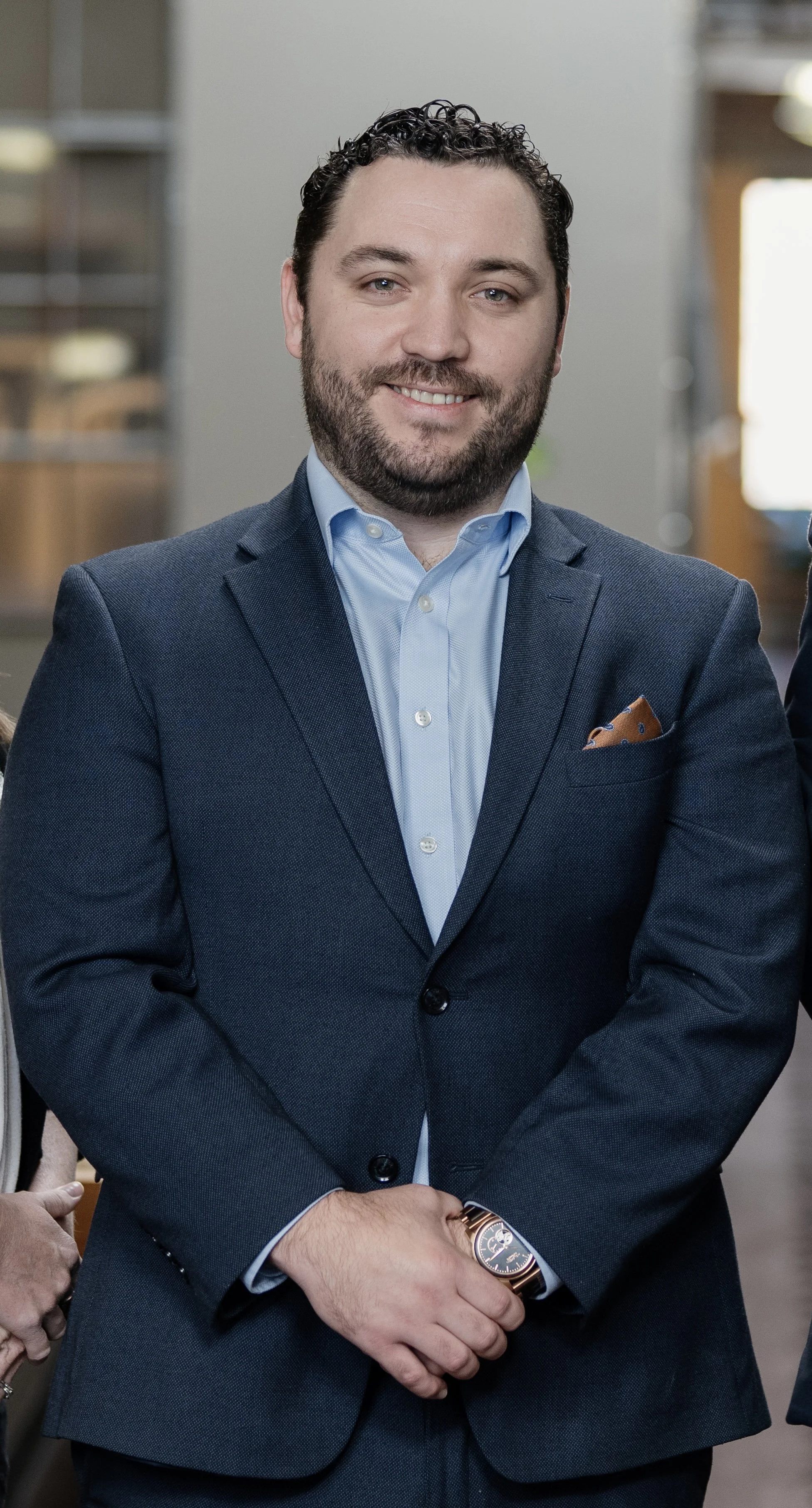 A man with dark curly hair and a beard, smiling in a blue suit and light blue shirt, standing indoors with his arms crossed, wearing a watch.
