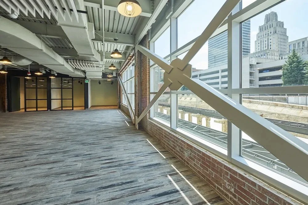 Empty modern interior space with wood flooring, exposed brick walls, large floor-to-ceiling windows, and a view of city buildings.