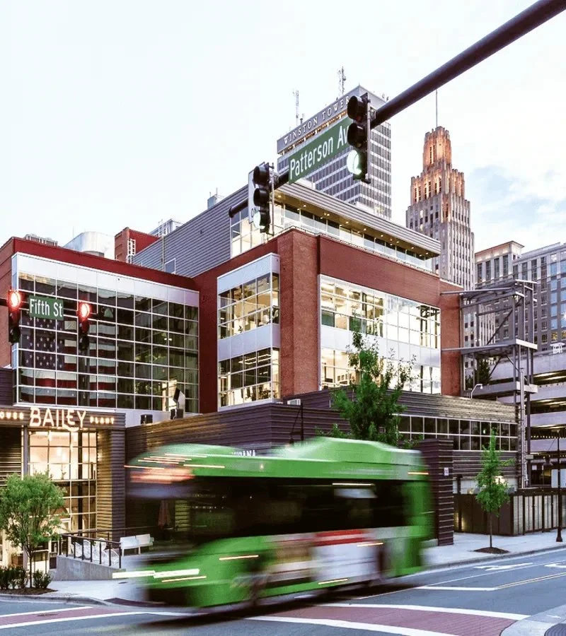 A city street scene with a moving green trolley, modern multi-story buildings, traffic lights, and street signs, including Fifth Street and Patterson Avenue, with a tall skyscraper in the background.