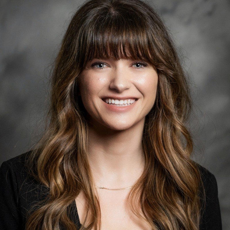 Portrait of a young woman with long, wavy brown hair and bangs, smiling and wearing a black top and a delicate necklace, against a gray background.