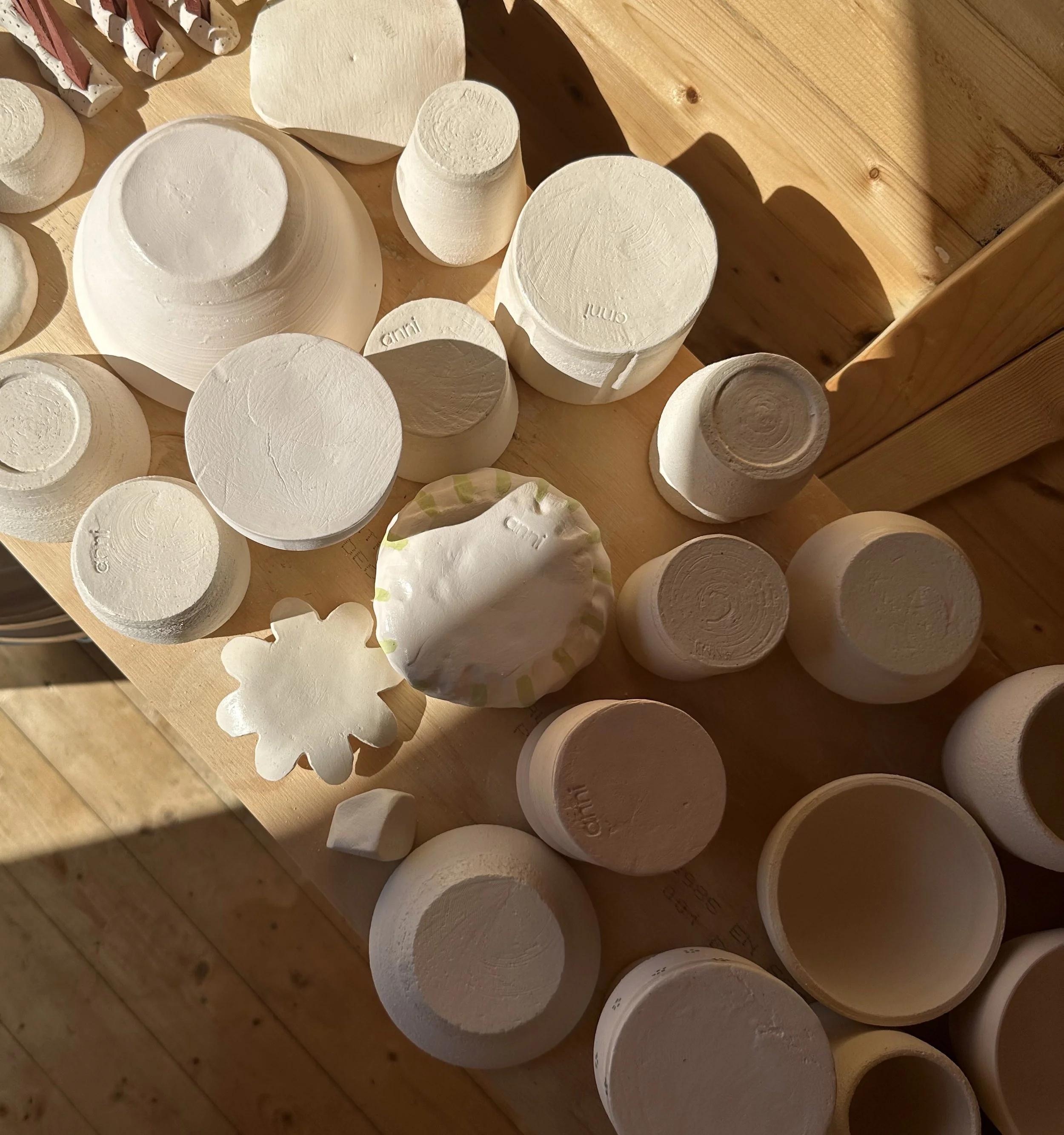 Various pottery pieces, including bowls and cups, in a raw, unglazed state on a wooden surface with sunlight casting shadows.