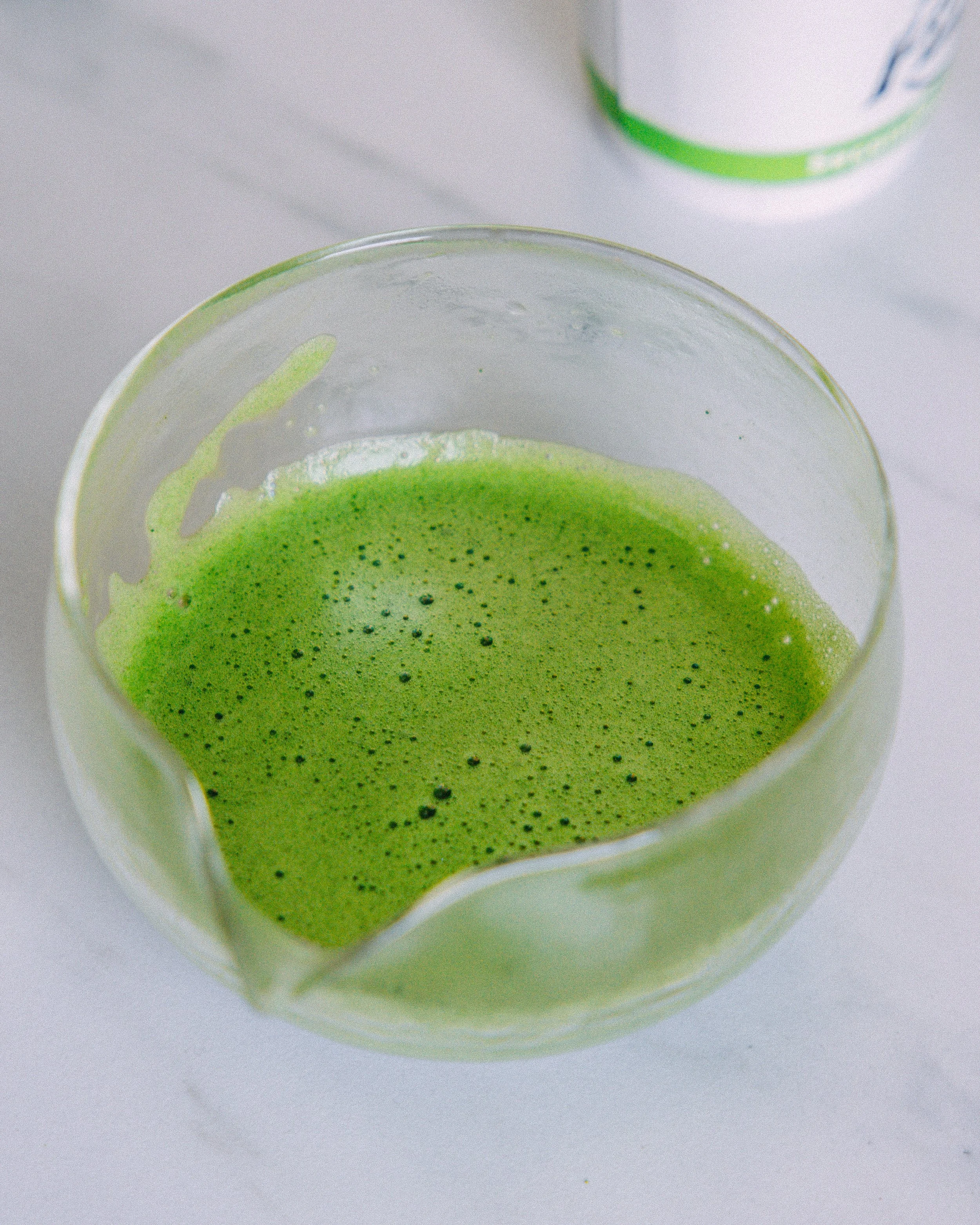 Close-up of a cup with green matcha tea, showing frothy surface with tiny dark green particles, and a separate container of matcha powder in the background.
