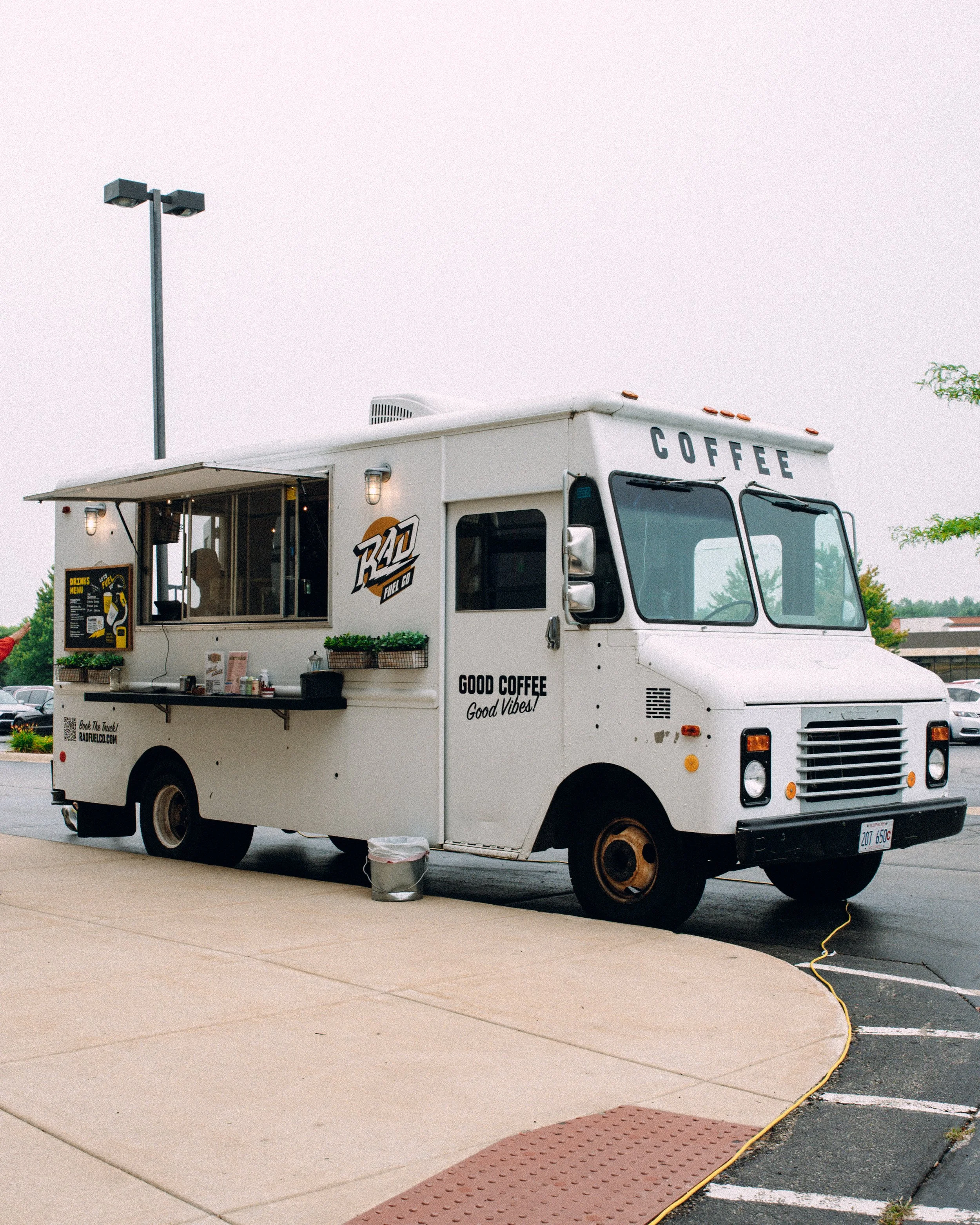 A white coffee food truck with the word 'COFFEE' displayed on the roof and a serving window open on the side. It is parked in a parking lot with a light pole and other vehicles visible in the background. There are signs and decorations on the truck, and an extension cord runs from the truck to an outlet.