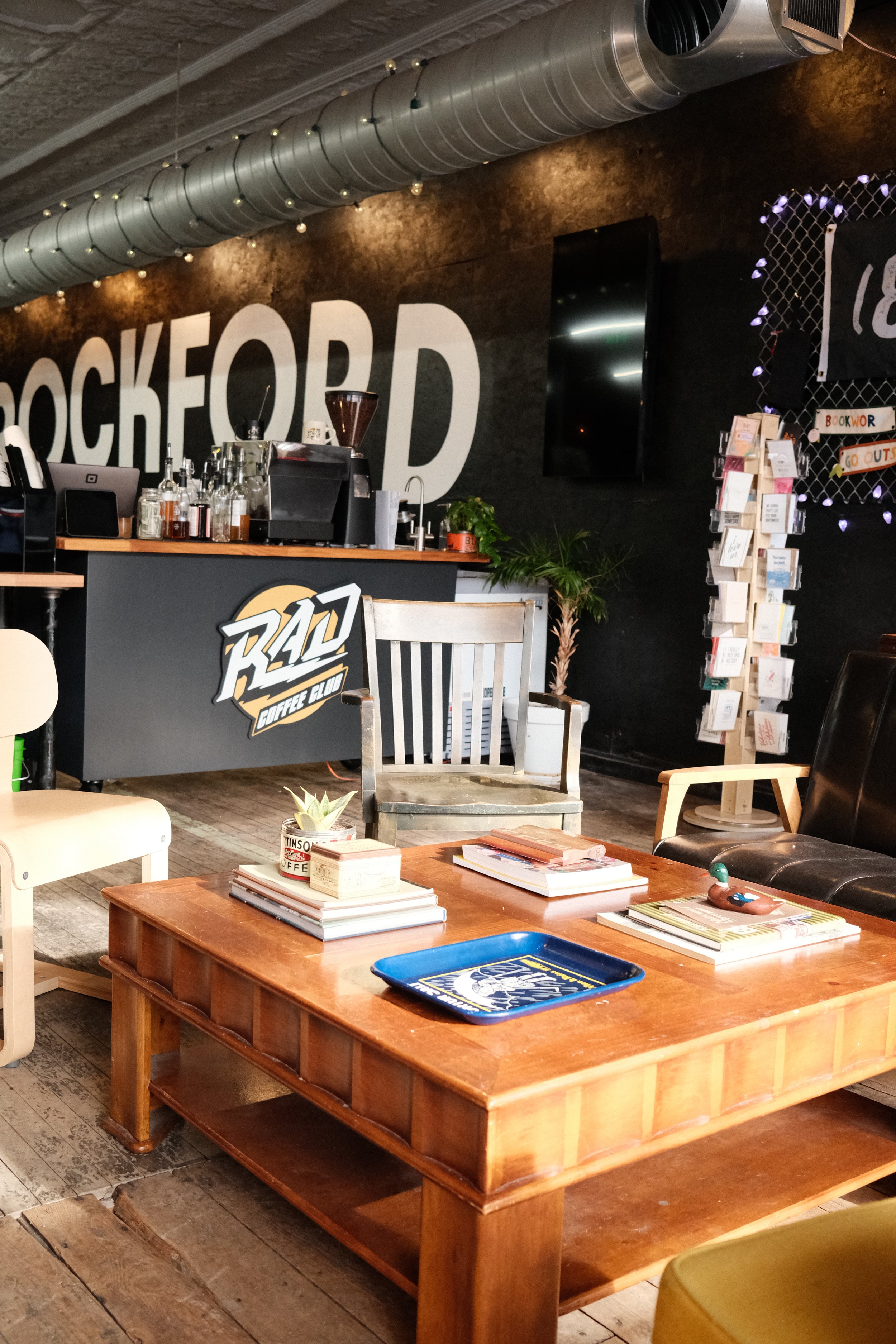 Rockford Coffee Club - Rockford Art Deli / Interior of a cozy coffee shop with a large black wall, a wooden table with books and a tray, a cafe counter with coffee equipment, and a reading display with postcards or greeting cards.