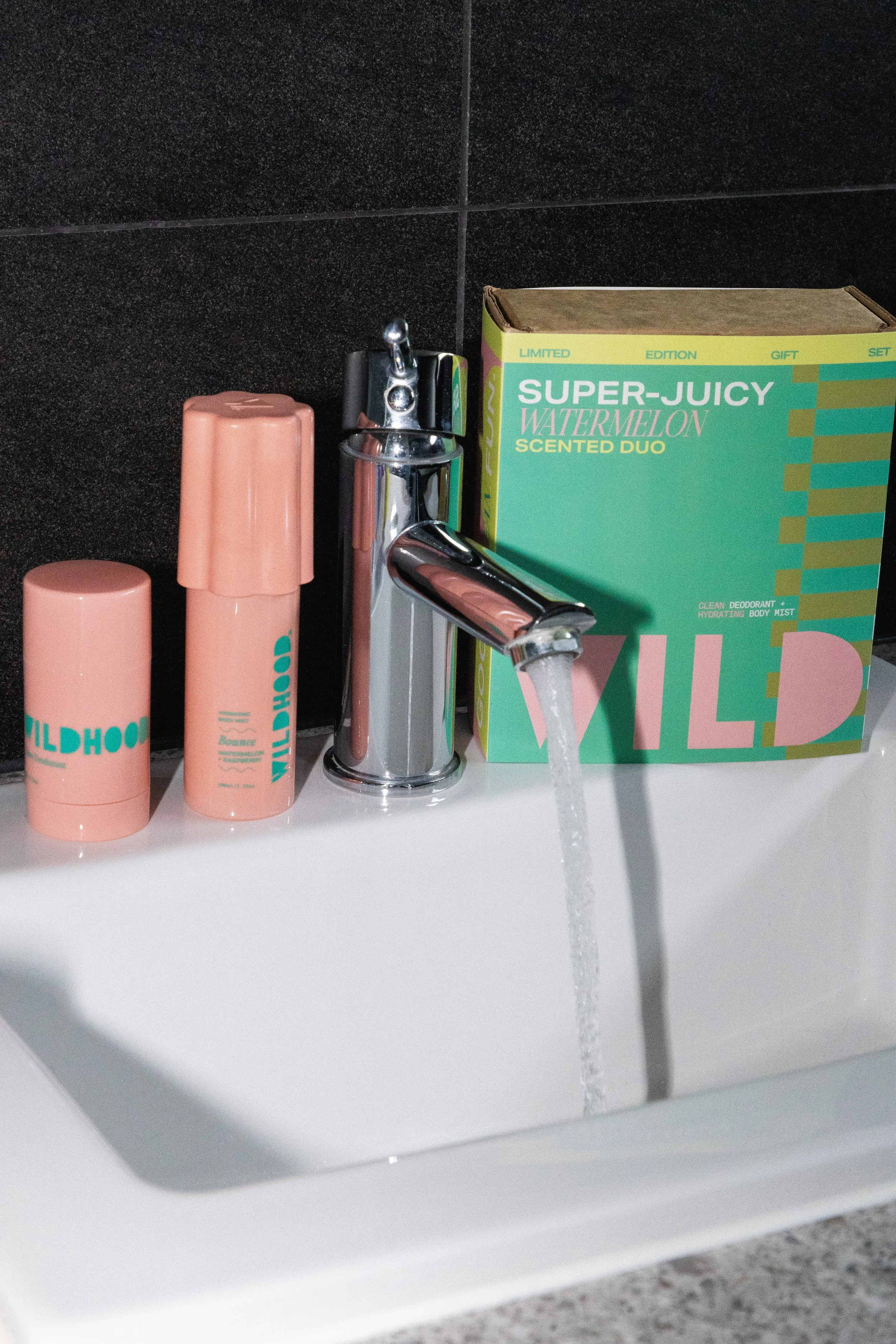 Bathroom sink with a silver faucet running water, containing skincare products in pink containers labeled "WILDHOO" and a green box of "Super-Juicy Watermelon Scented Duo" body mist, against a black tiled wall.